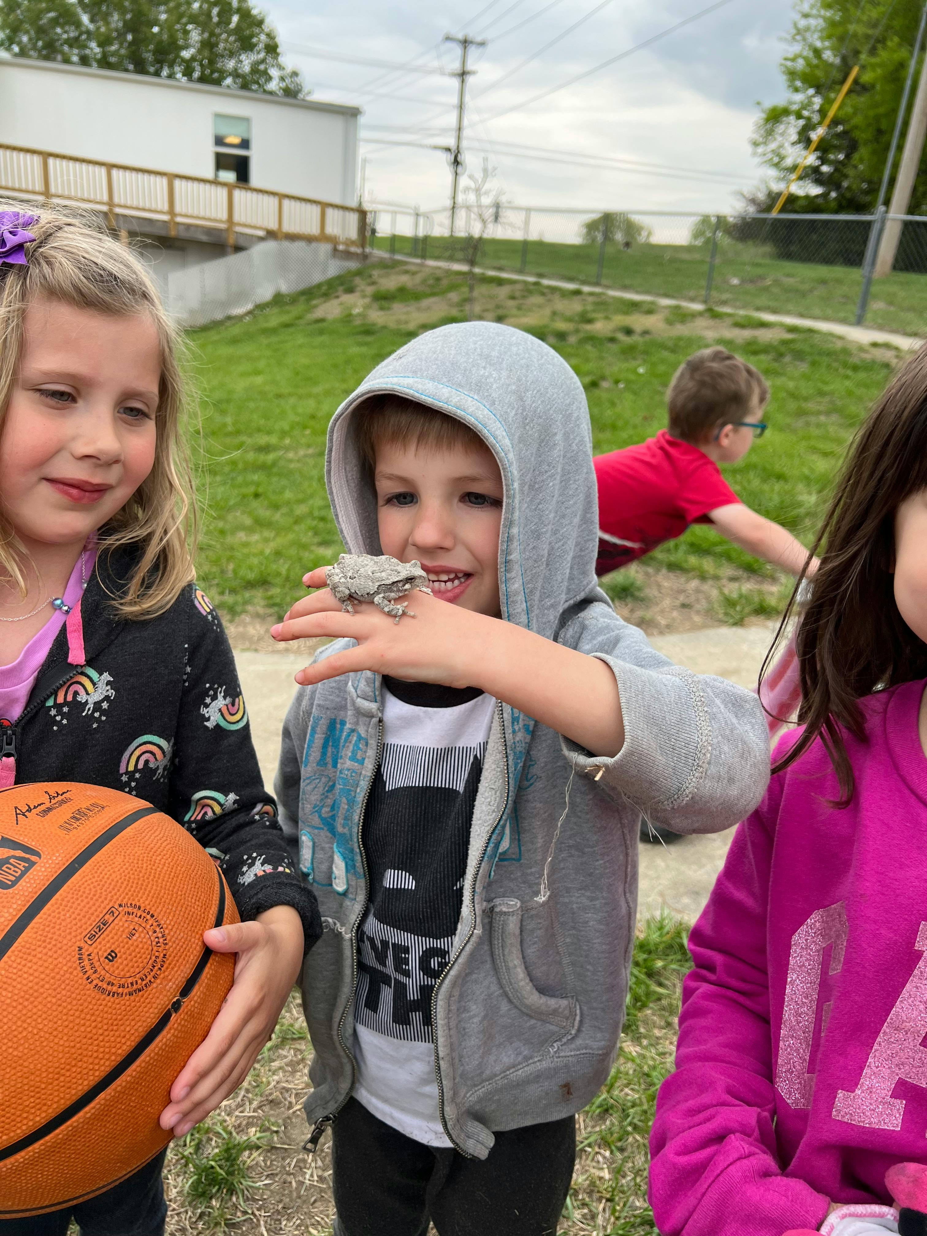 Child in a gray hoodie holding a frog on their hand, with other children nearby