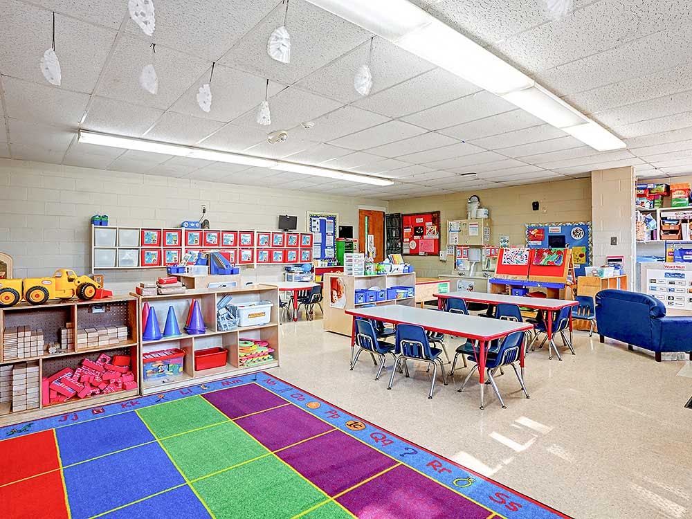 Classroom with small tables and chairs, shelves with toys, and an alphabet rug