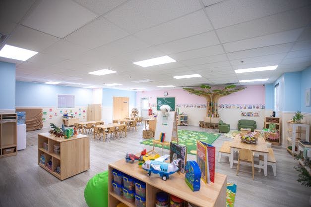 Empty classroom with small tables and chairs, shelves with toys, and a tree mural