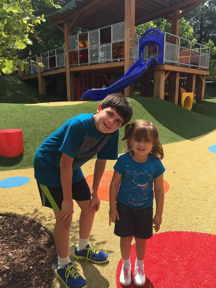 Two children standing on a playground with a large play structure in the background