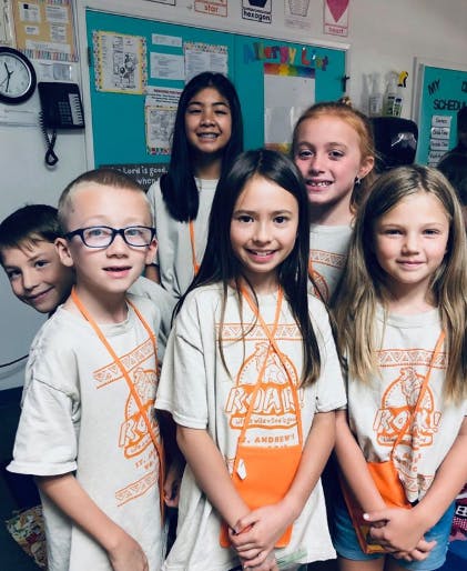 Six children in a classroom, some wearing t-shirts with a logo and orange pouches