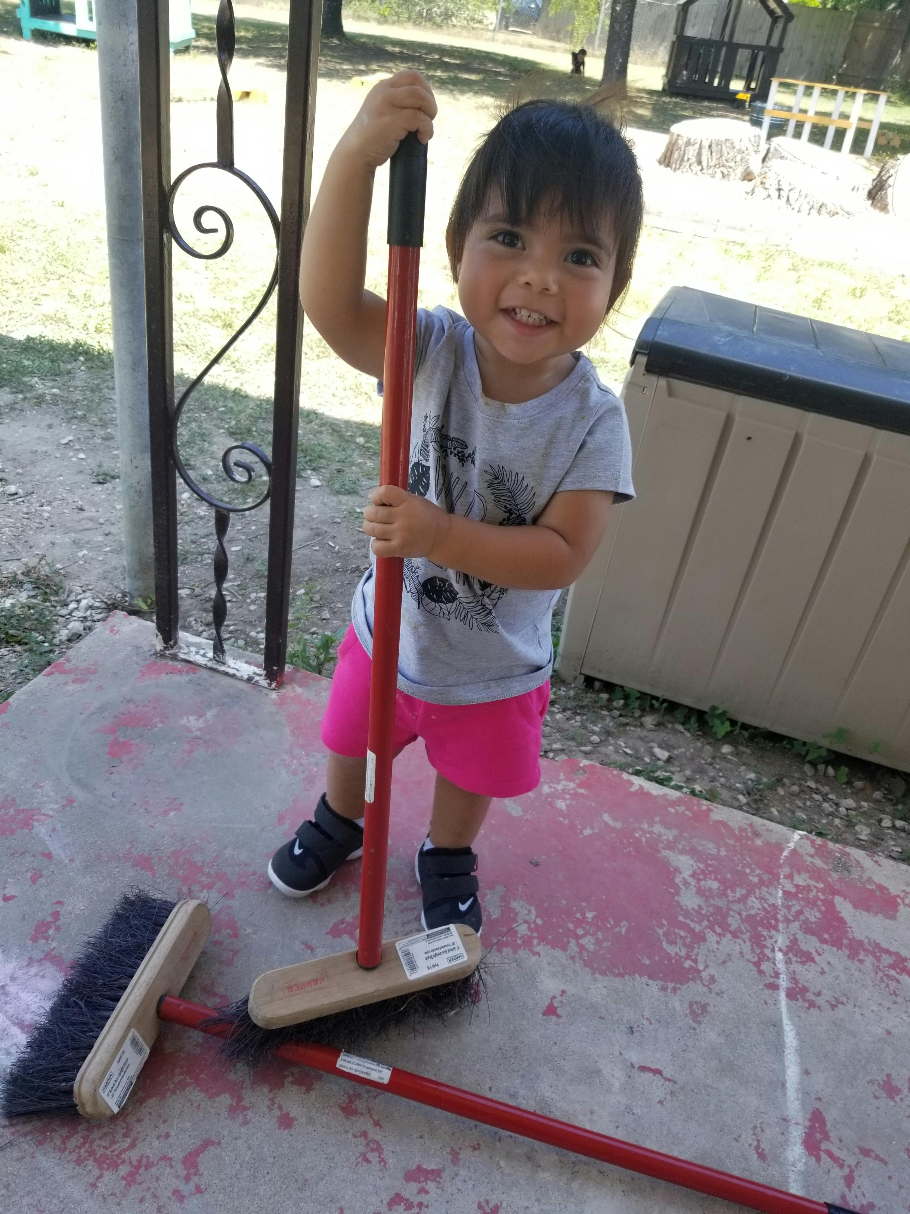 Child holding a red broom, with two other brooms on the ground