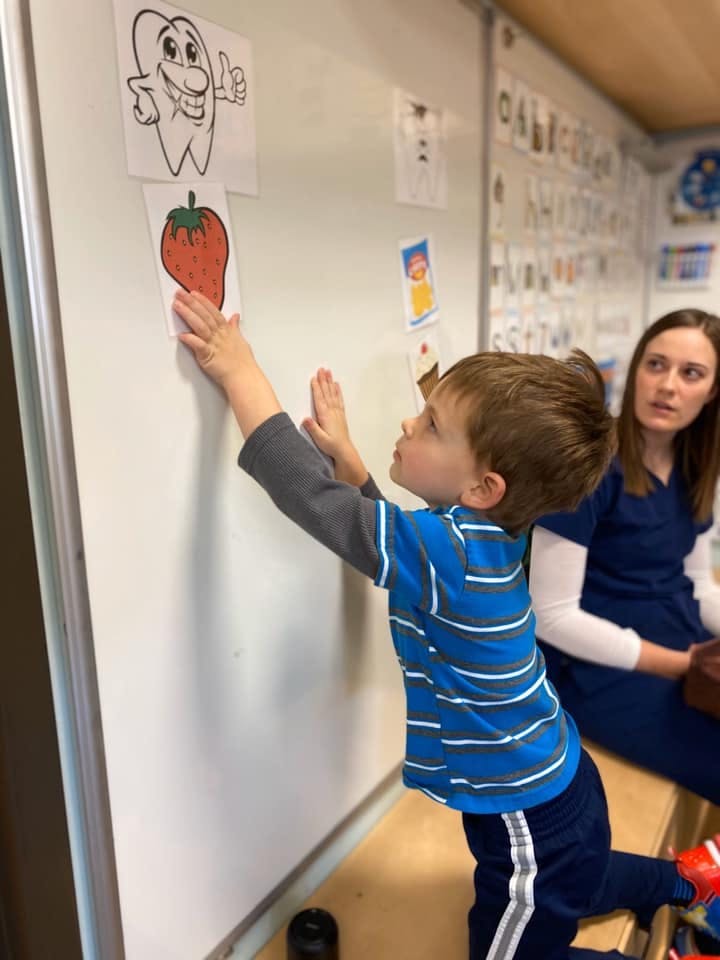 Child reaching for a strawberry drawing on a whiteboard, person in background
