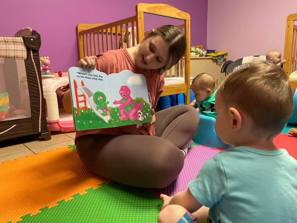 Person reading a book to a child in a room with other children in the background