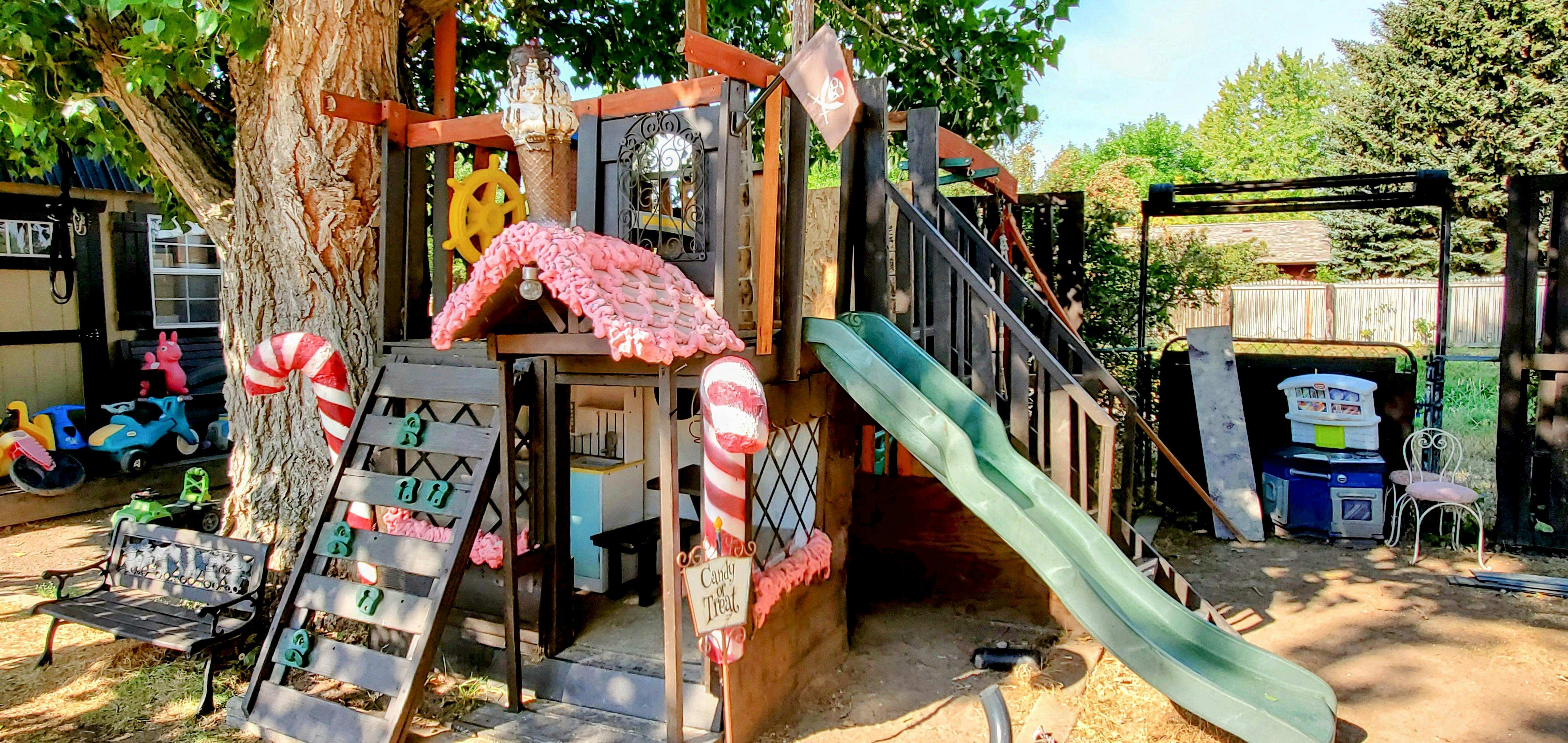 A wooden playset with a green slide attached to a large tree in a backyard