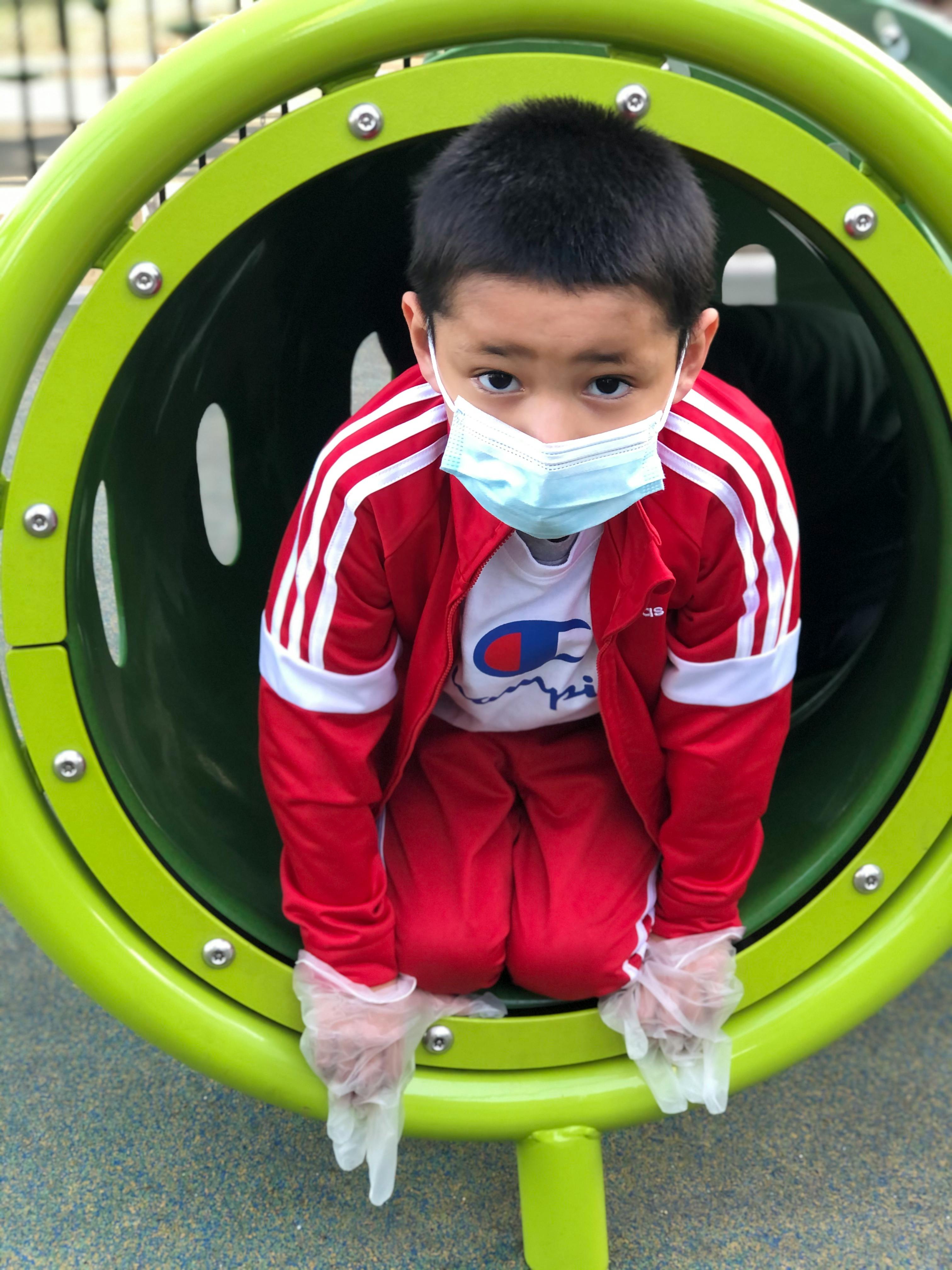 Child in a face mask and gloves kneeling in a green playground tunnel