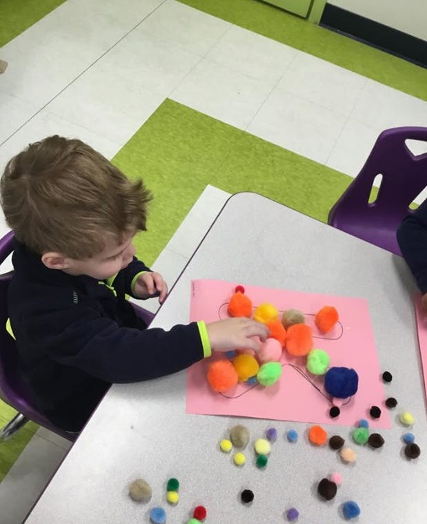 Child playing with colorful pom-poms on pink paper at a table