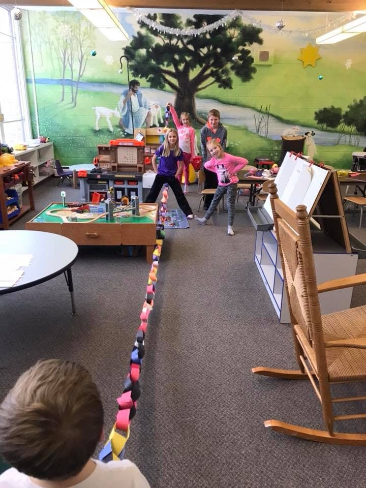 Four children standing in a classroom with a long paper chain on the floor