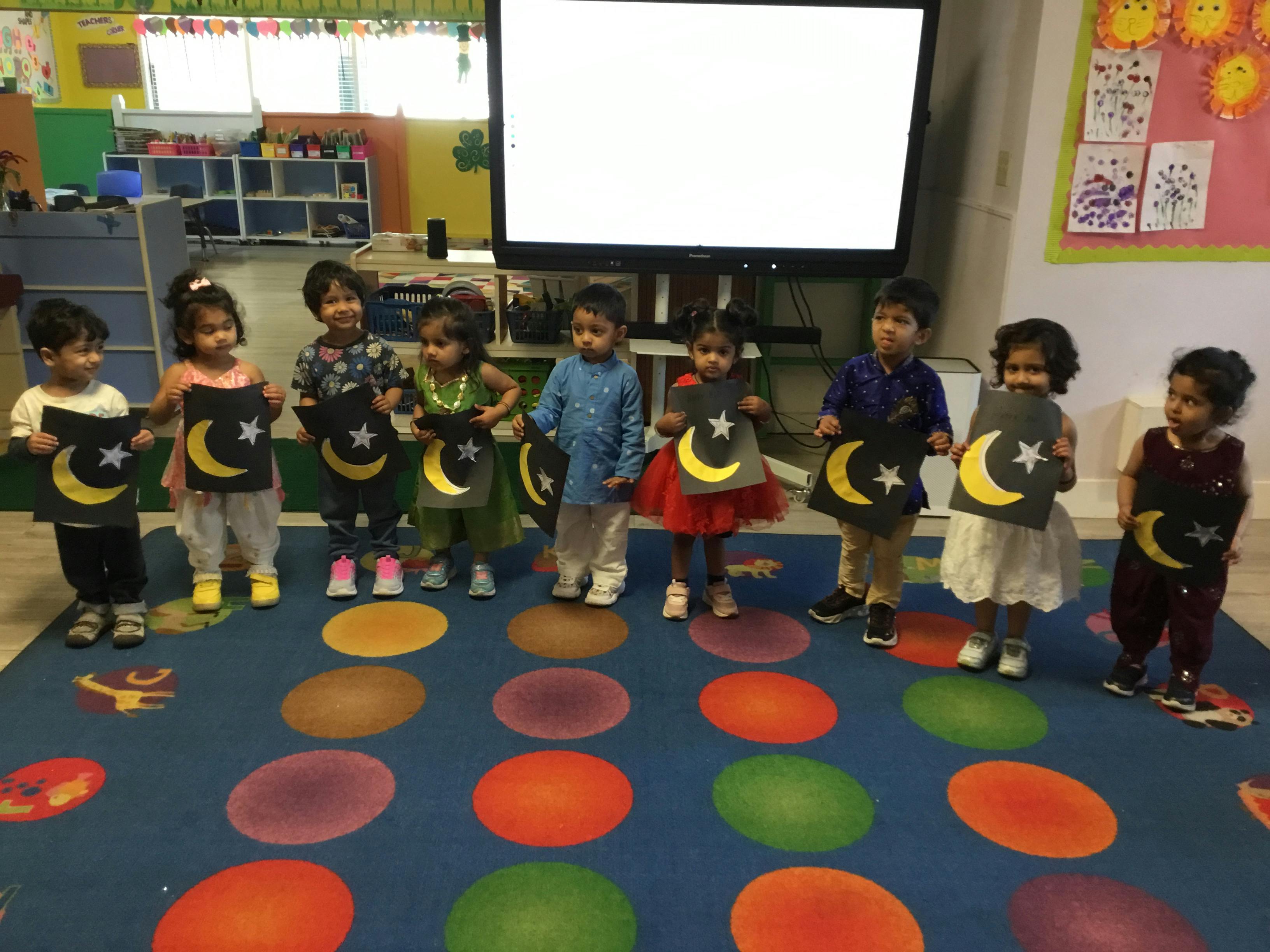 Eight children in a classroom holding up crescent moon and star crafts