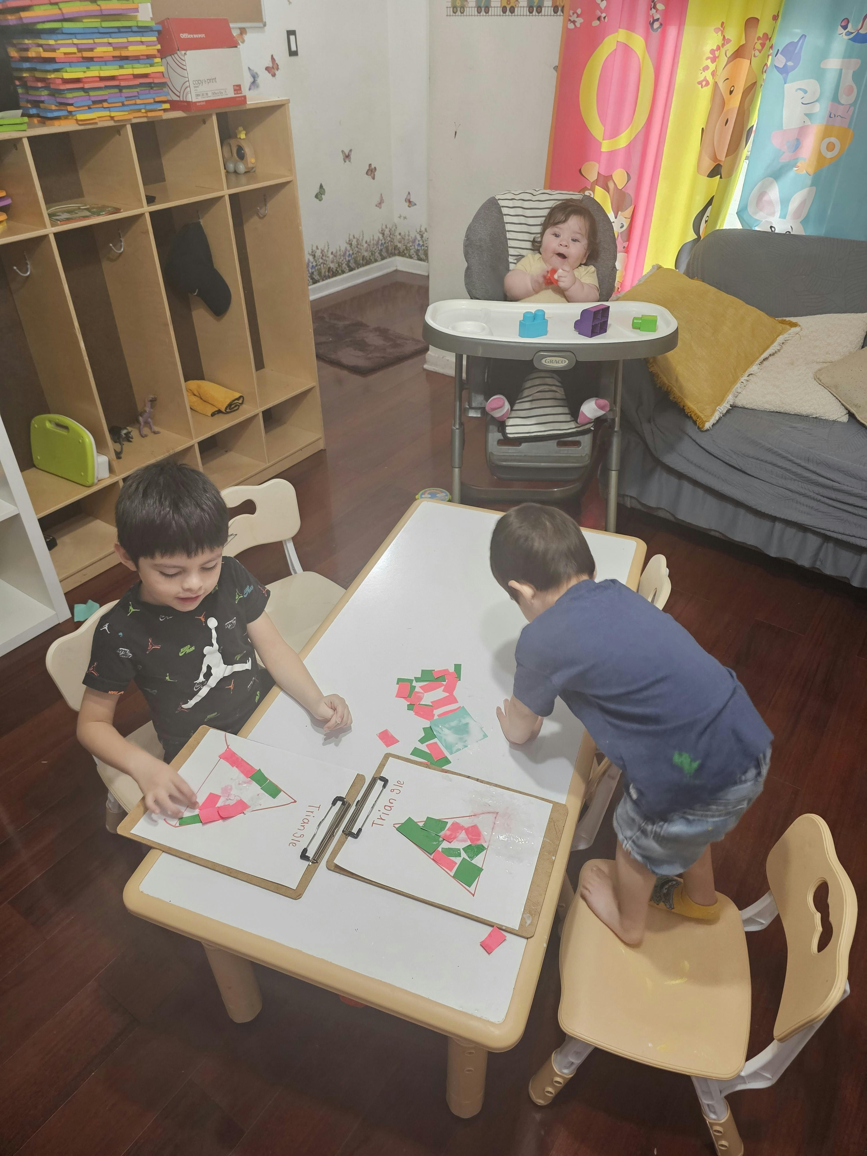 Two children at a table with paper and shapes, a baby in a high chair