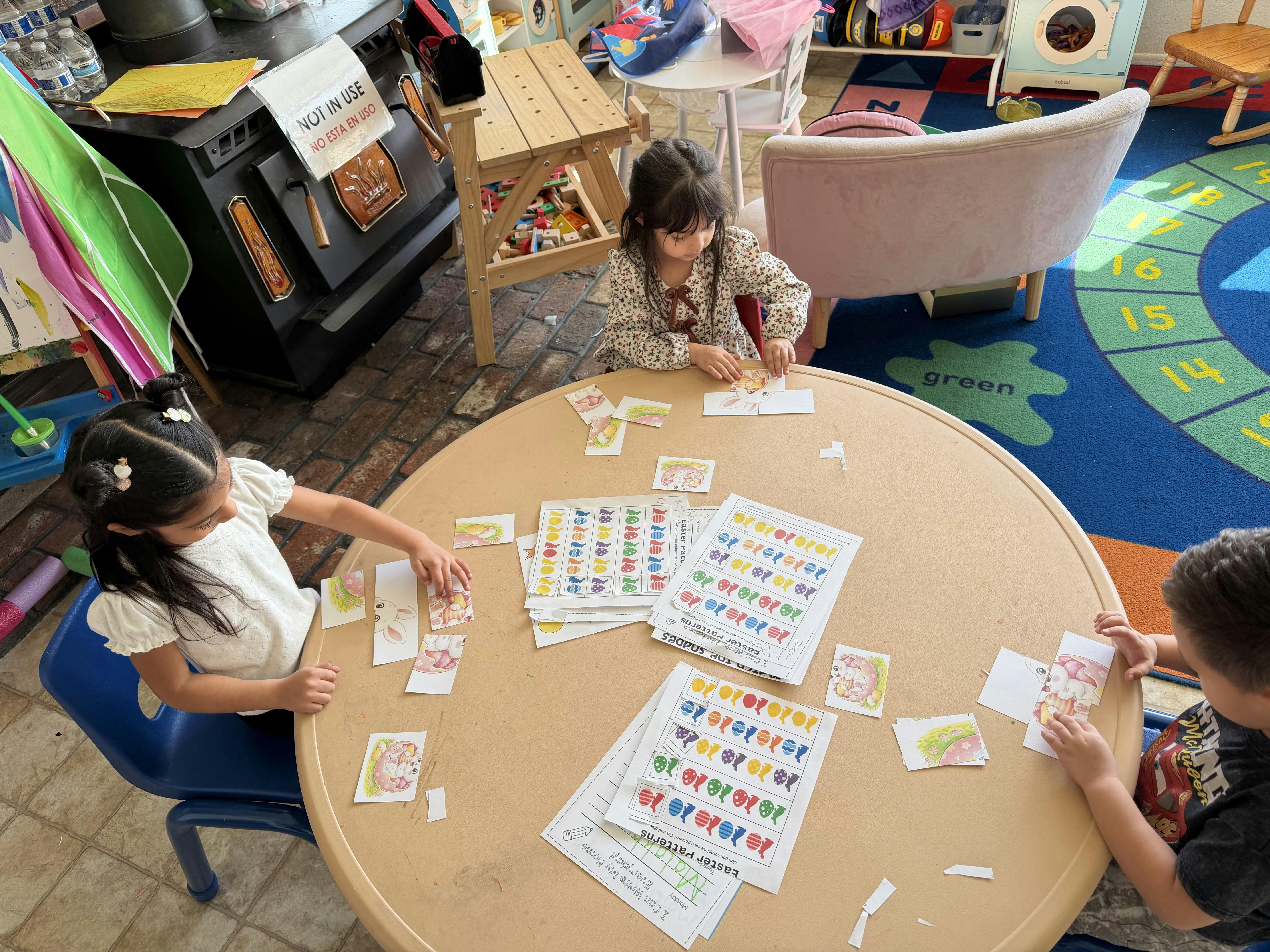 Three children sitting around a table with paper cutouts and worksheets