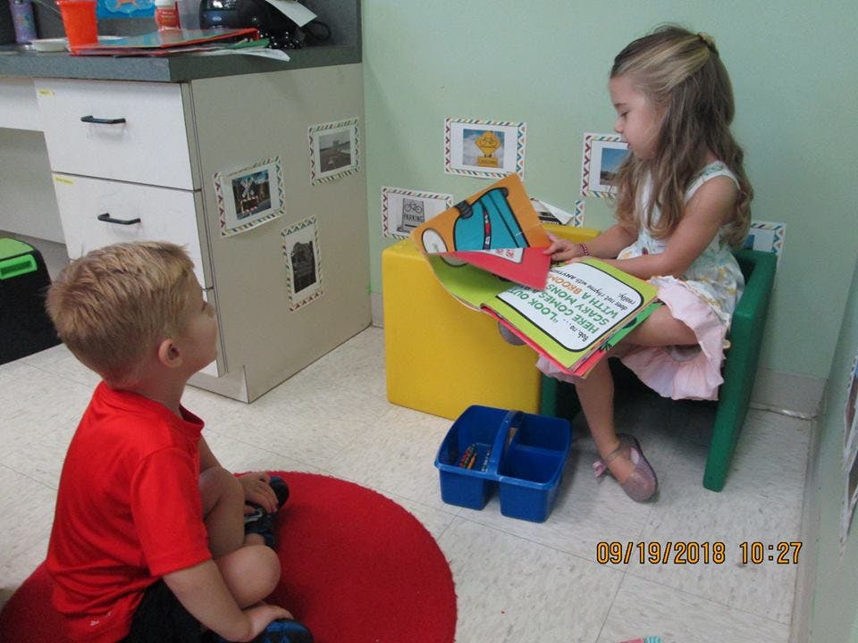 Child sitting on floor looking at child reading a book