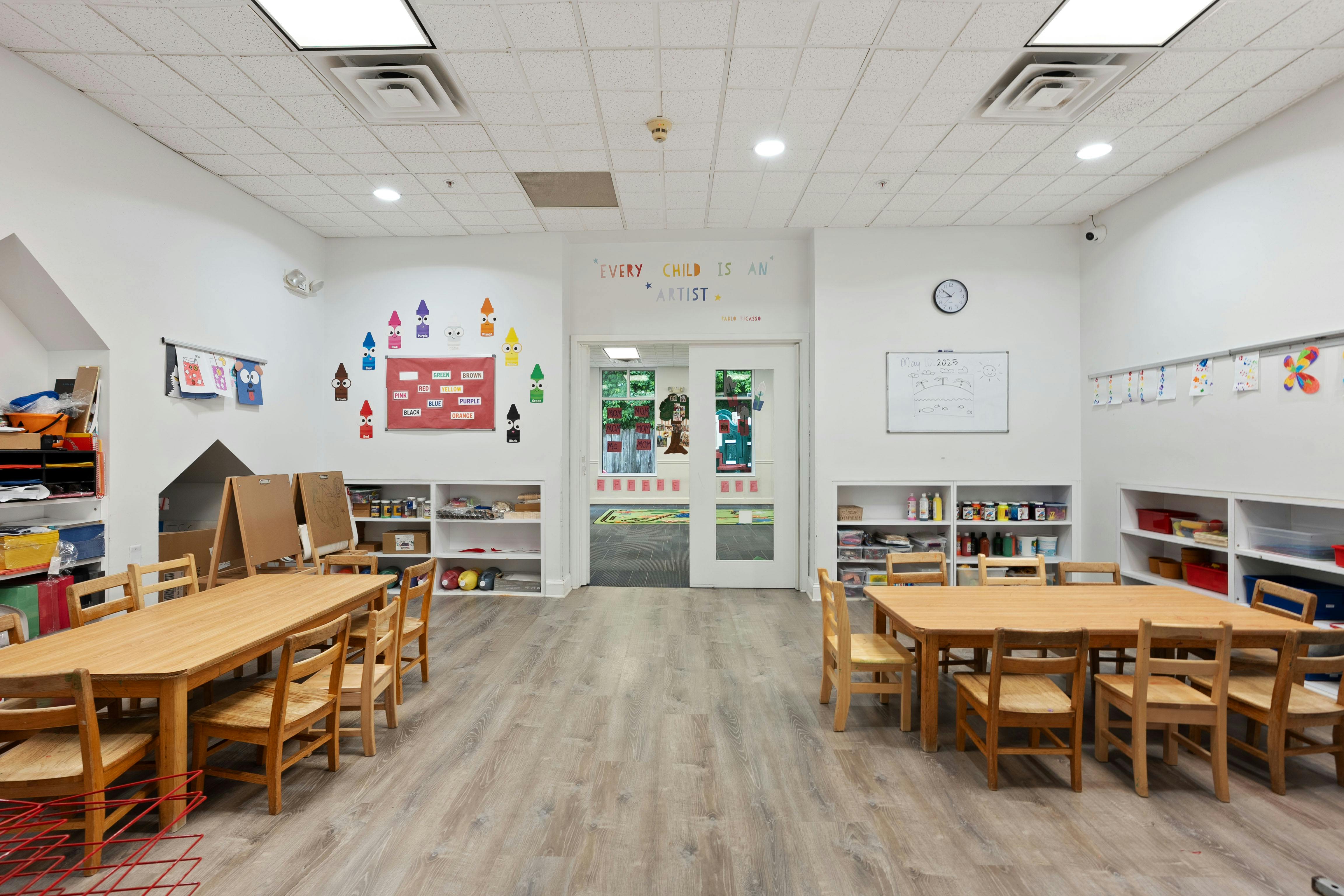 Classroom with wooden tables and chairs, art supplies on shelves, and a whiteboard