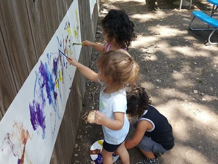 Three children painting on a long sheet of paper attached to a wooden fence