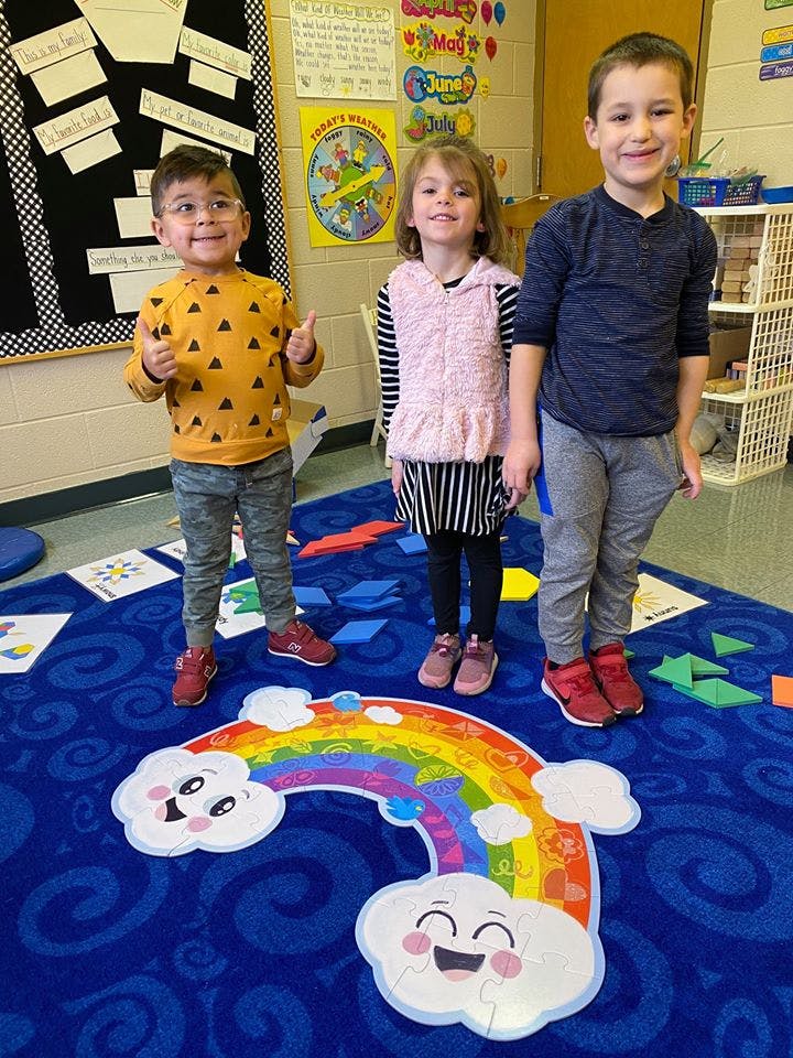 Three children standing on a blue rug with a rainbow puzzle in a classroom