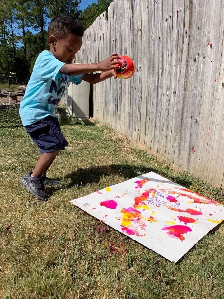 Child holding a colorful ball over a paint-splattered canvas on the ground