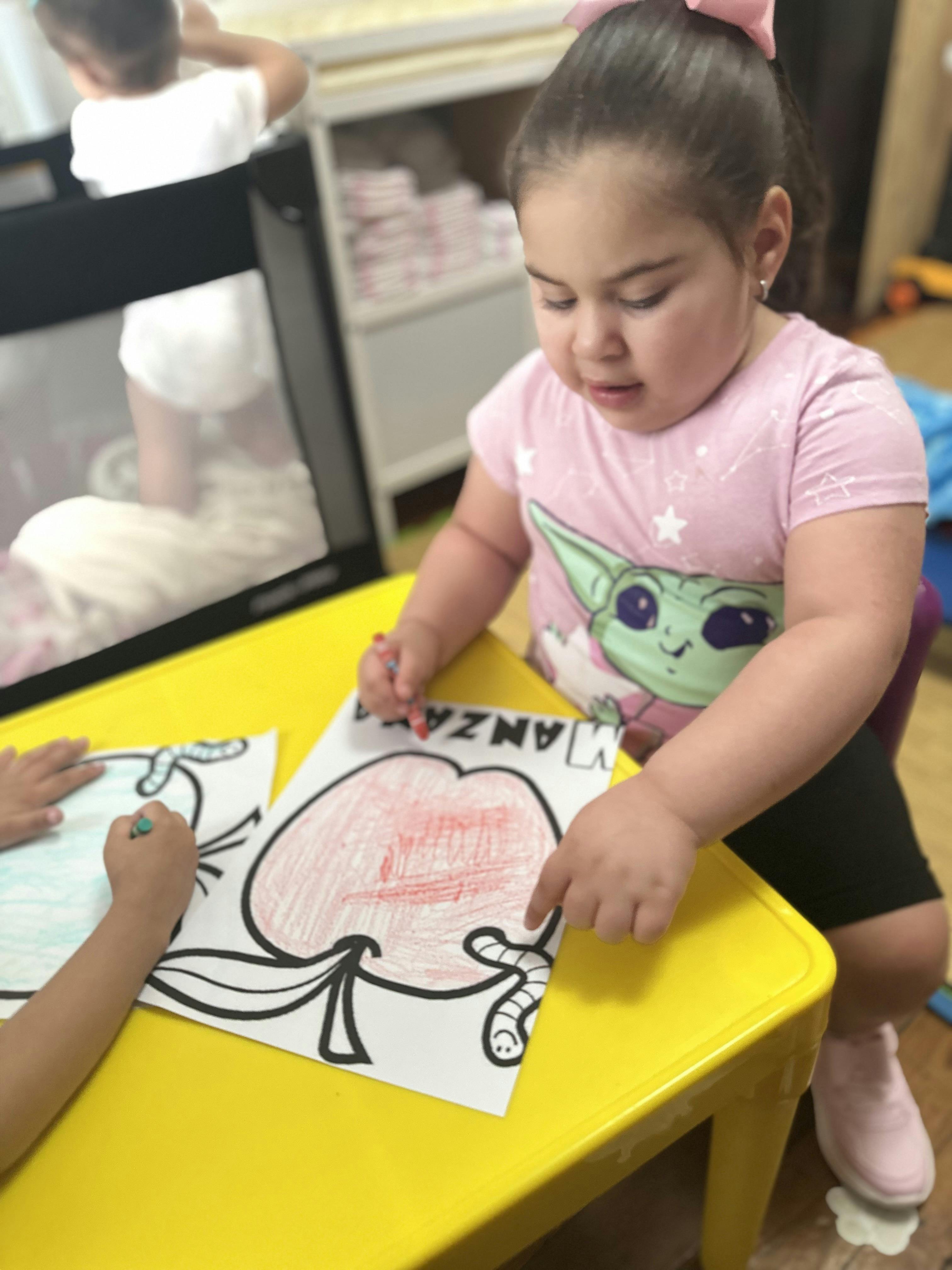 Child coloring a drawing of an apple with a worm on a yellow table