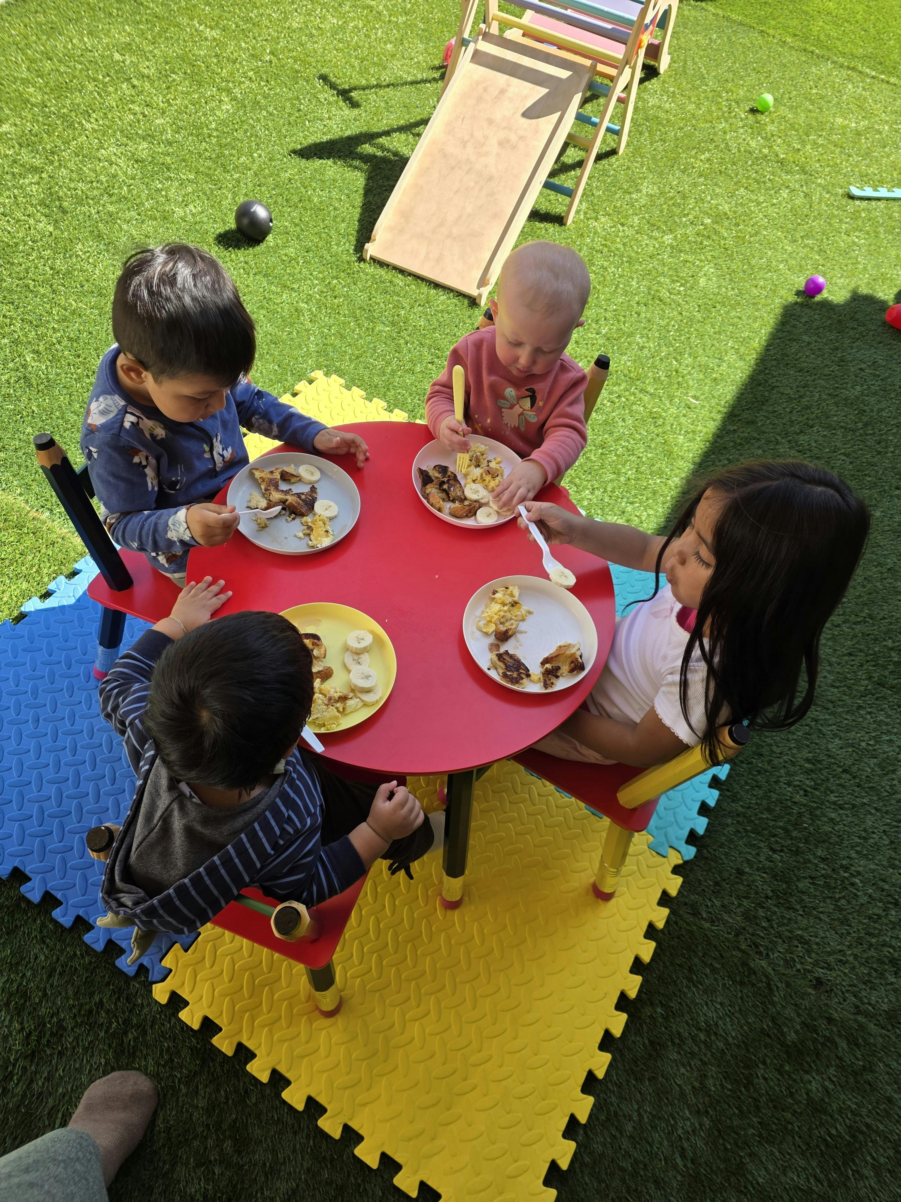Four children eating breakfast at a red table on a green artificial turf