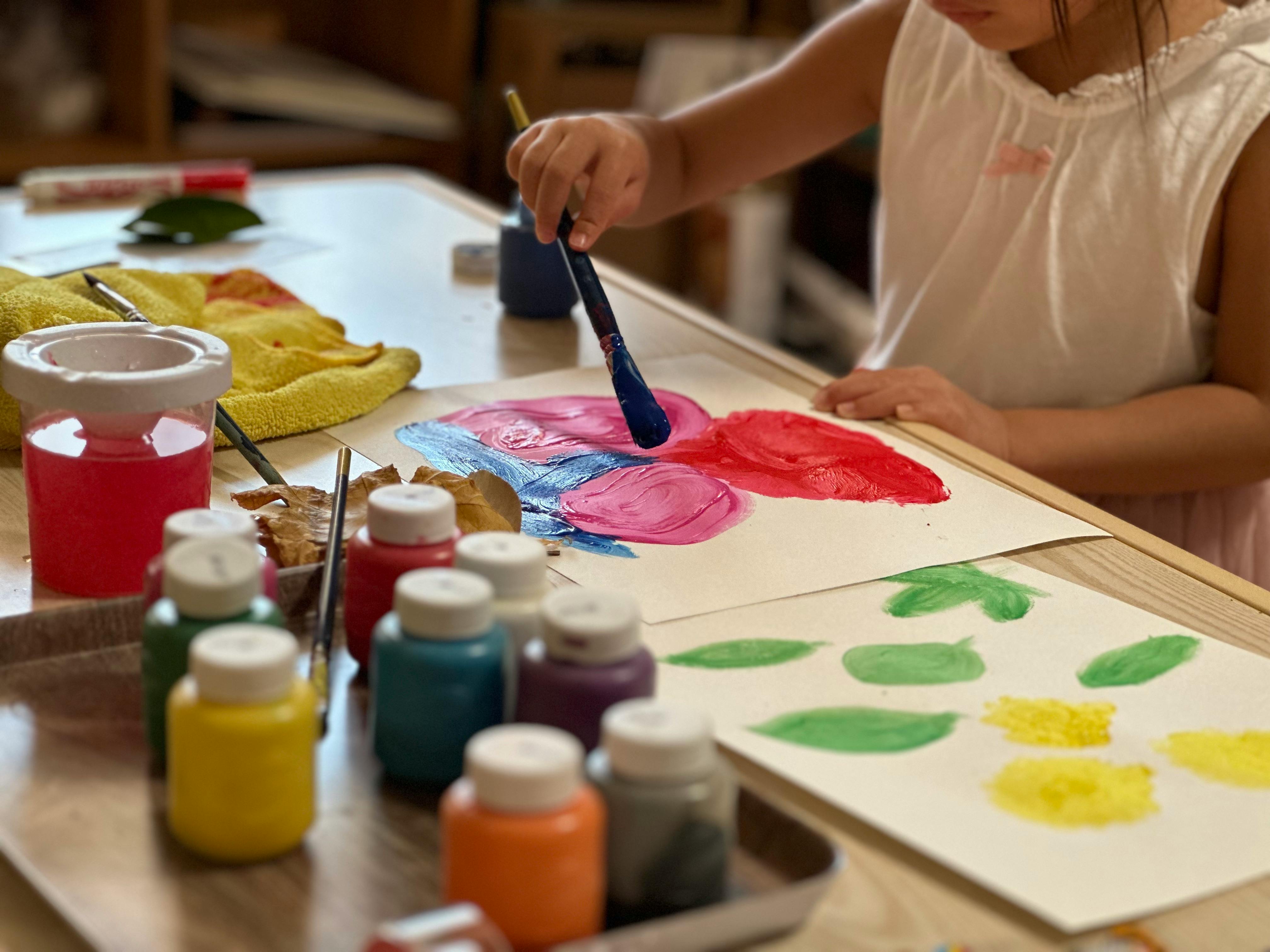 Child painting with a brush on paper, surrounded by paint bottles