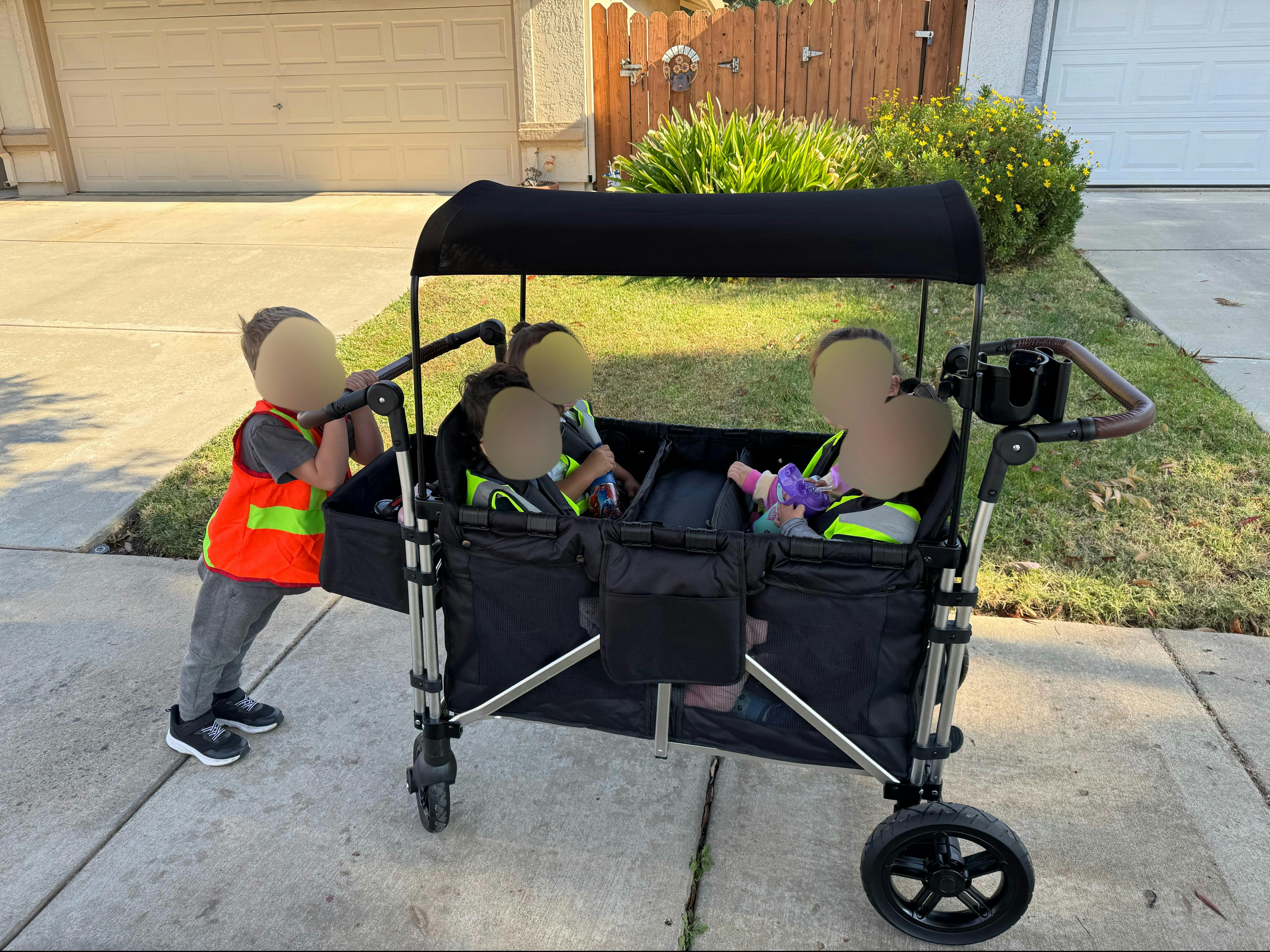 Child in orange vest pushing a wagon with three children in yellow vests
