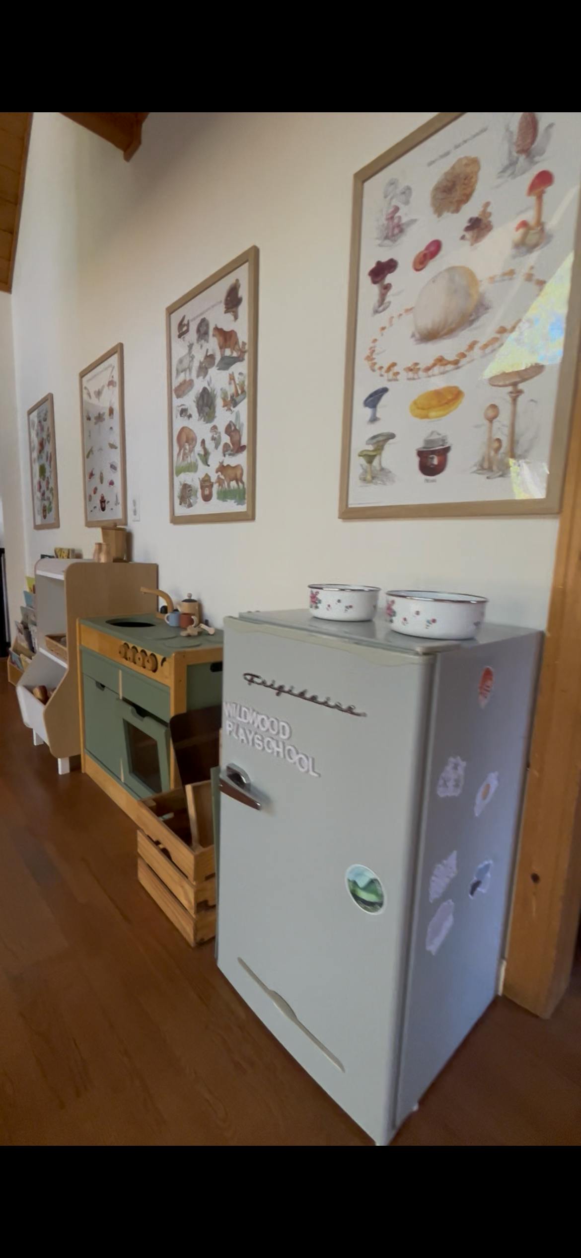 A grey toy refrigerator with the words "Wildwood Playschool" on its door, next to a toy kitchen