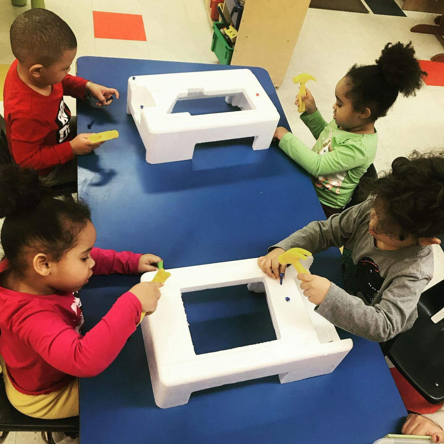 Four children playing with toy hammers and white plastic objects at a blue table