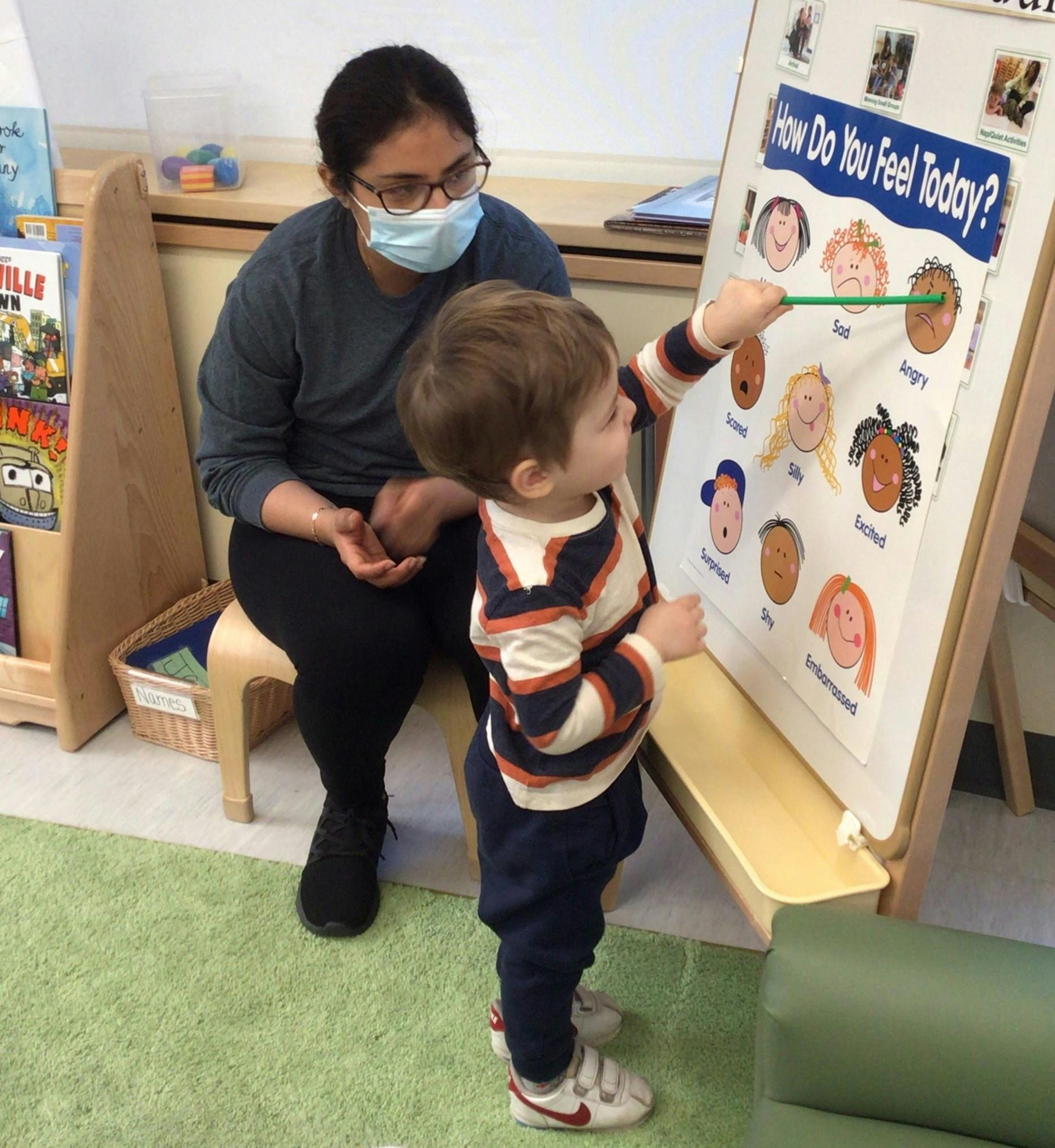 Person in mask watching child point to a feeling chart in a classroom