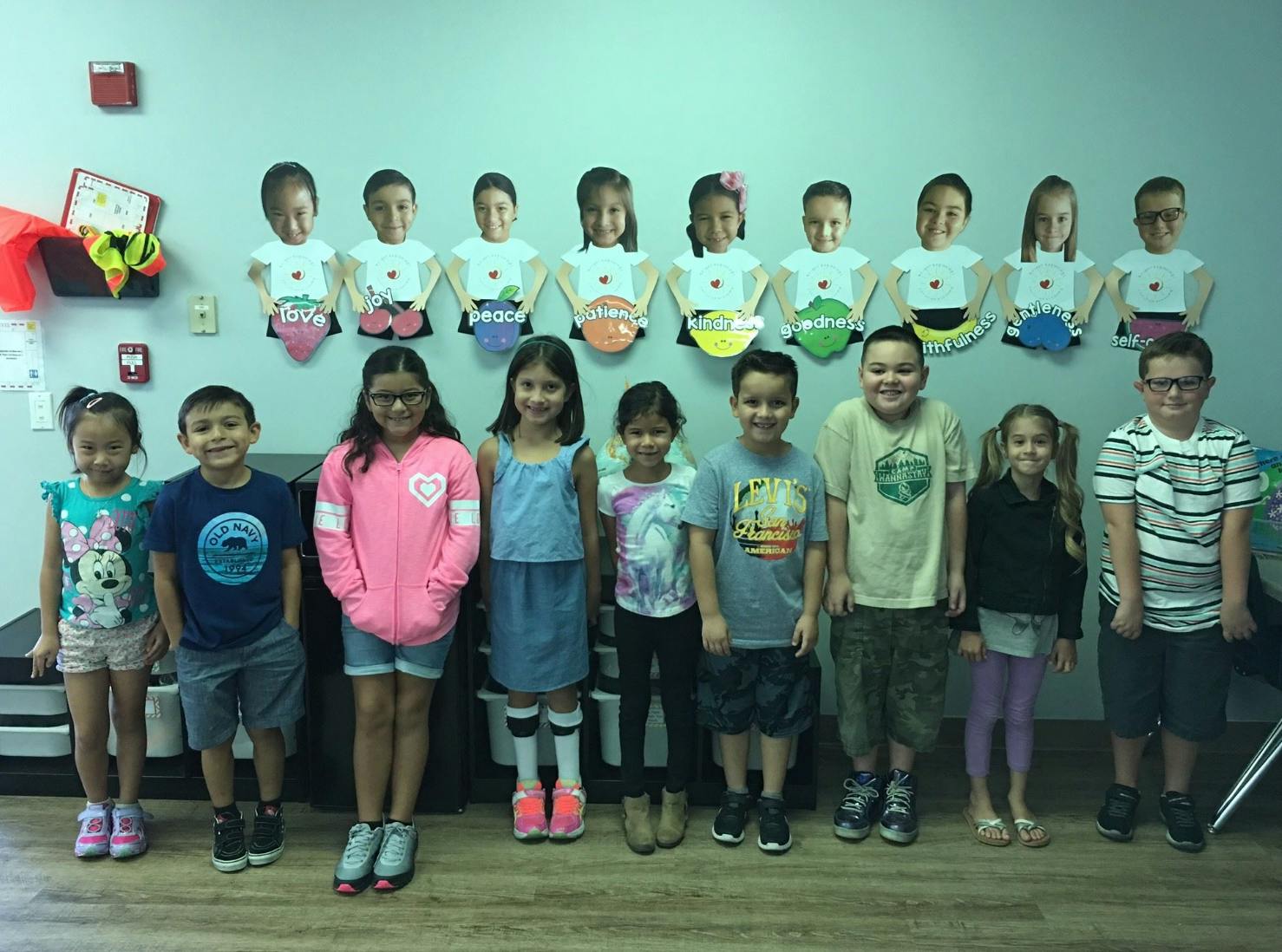 Group of children standing in a classroom with paper cutouts of children on the wall