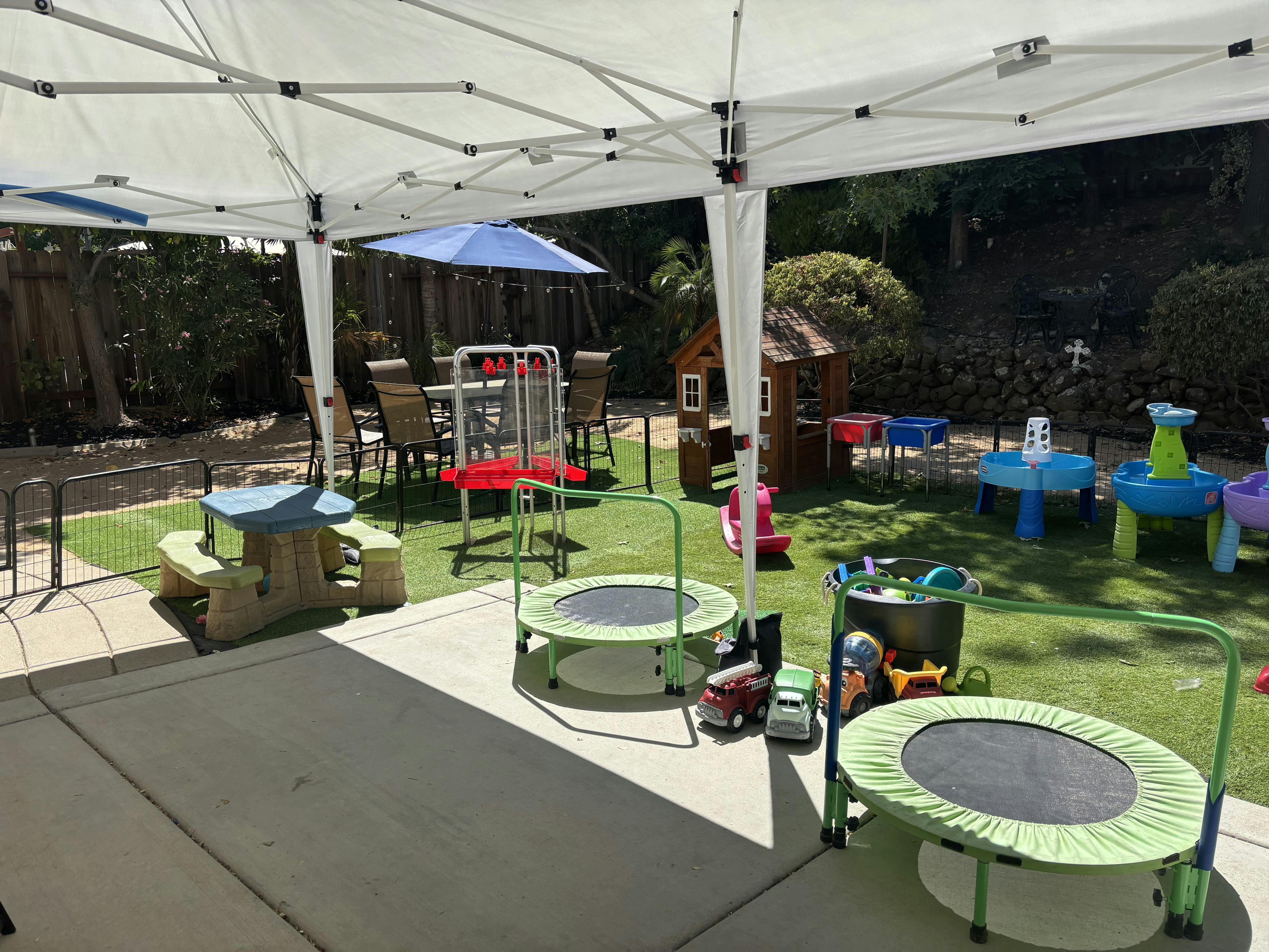 Outdoor play area with trampolines, a picnic table, and other toys under a tent