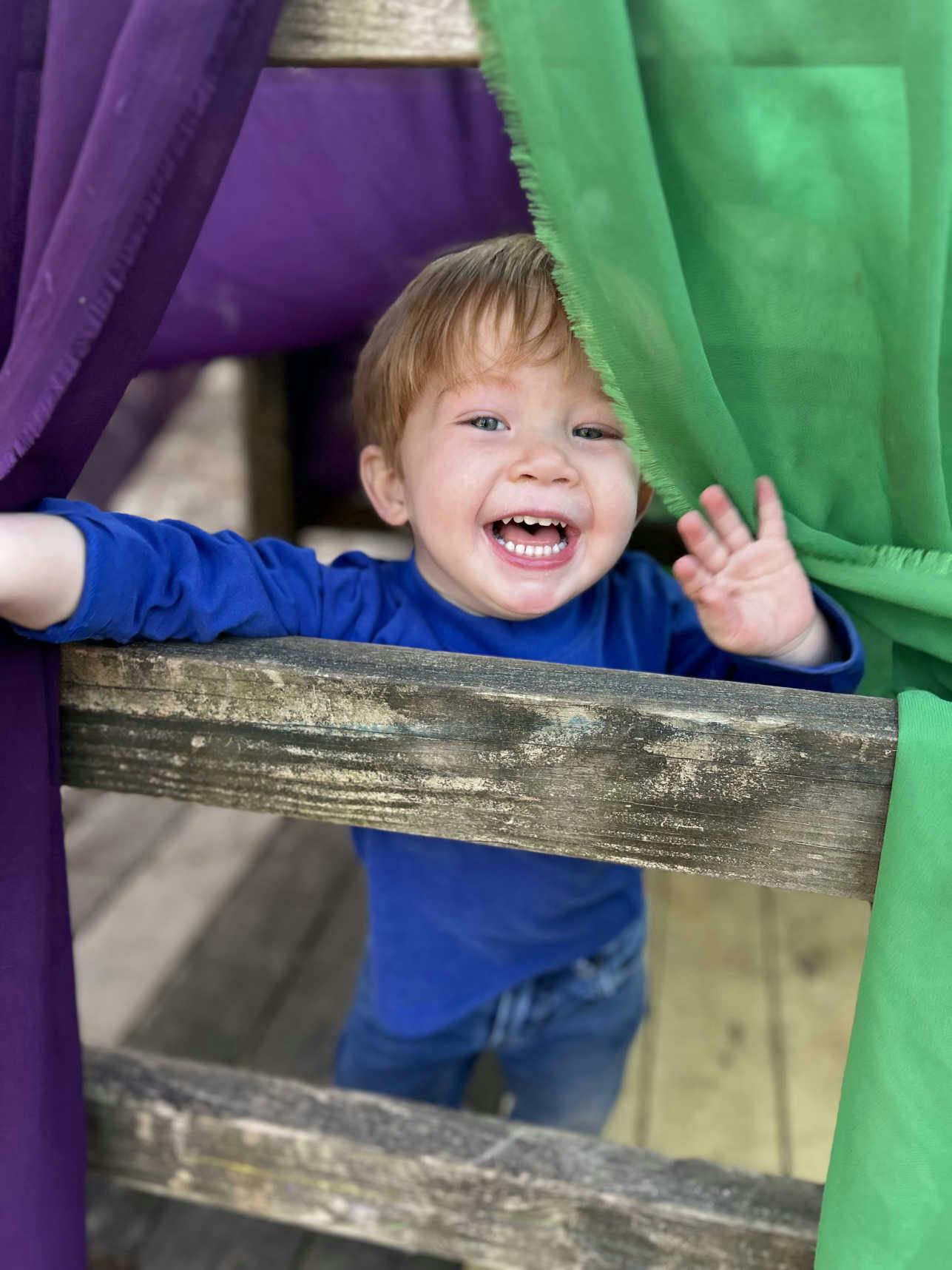 Child smiling and waving behind a wooden railing with purple and green fabric