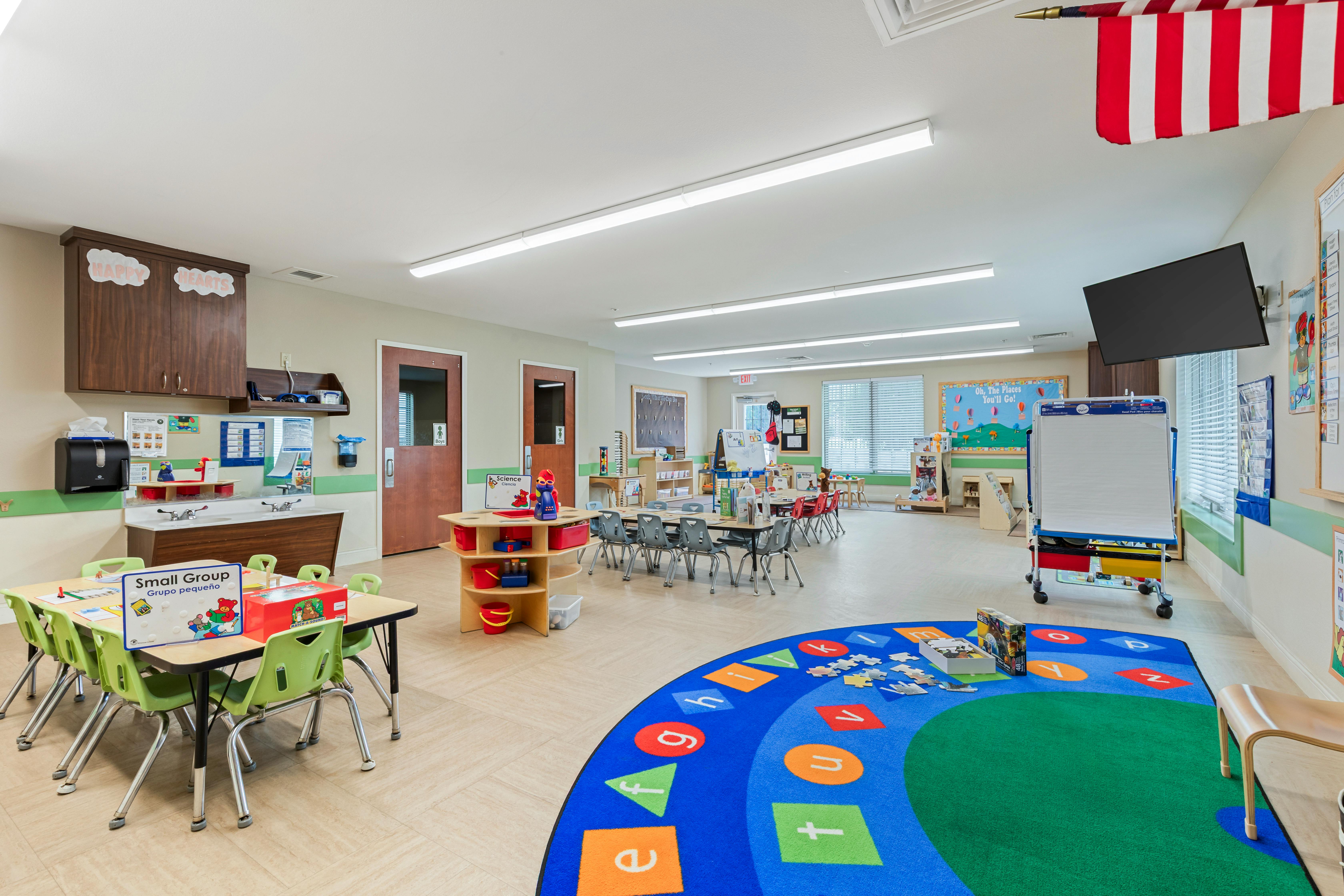 Classroom with small tables and chairs, a sink area, and a colorful rug