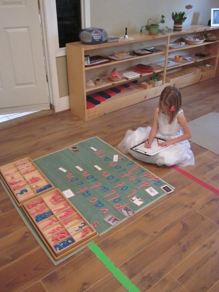 Child writing on paper next to a large alphabet mat on the floor