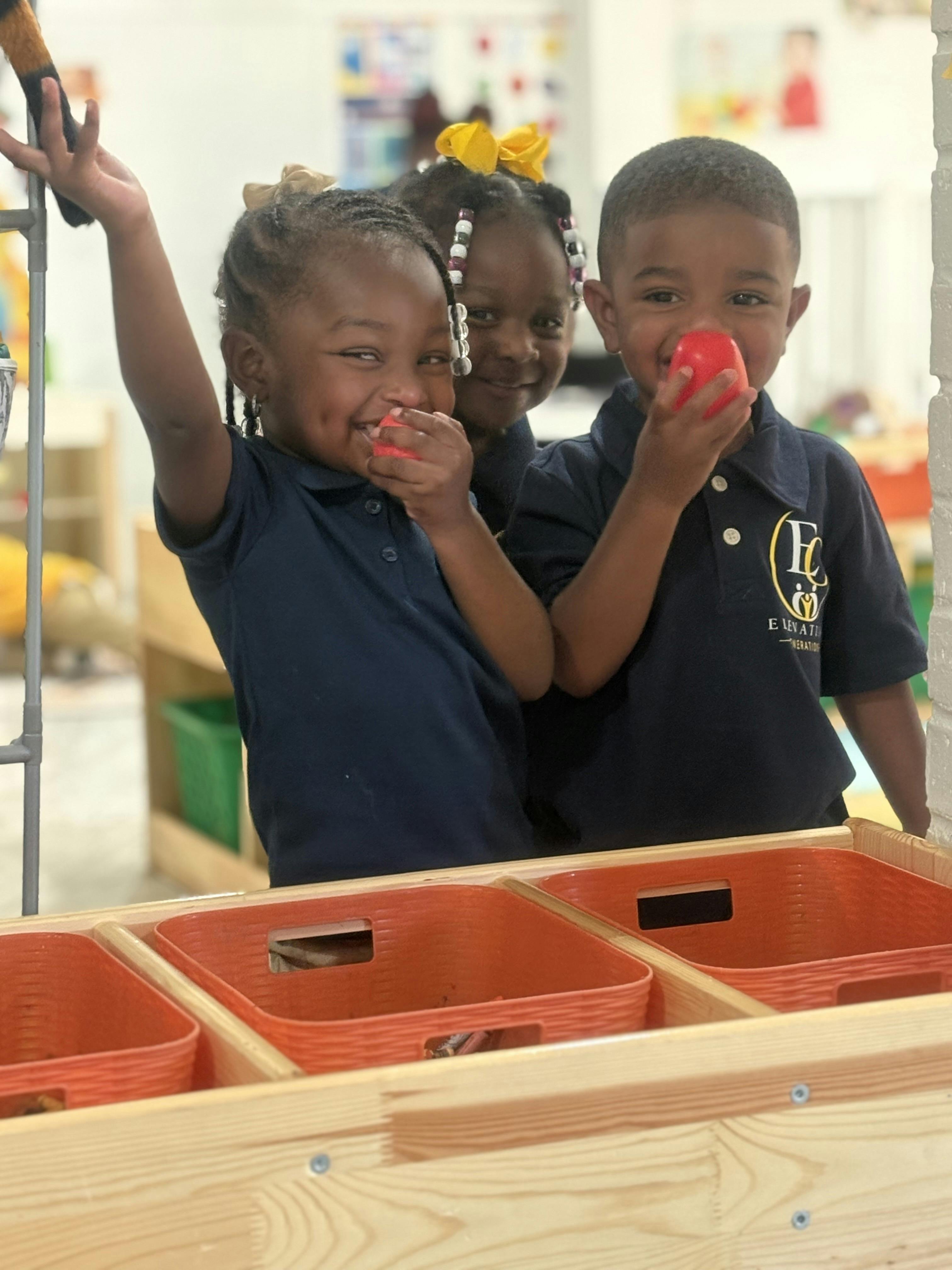 Three children in matching uniforms holding red play food in a classroom