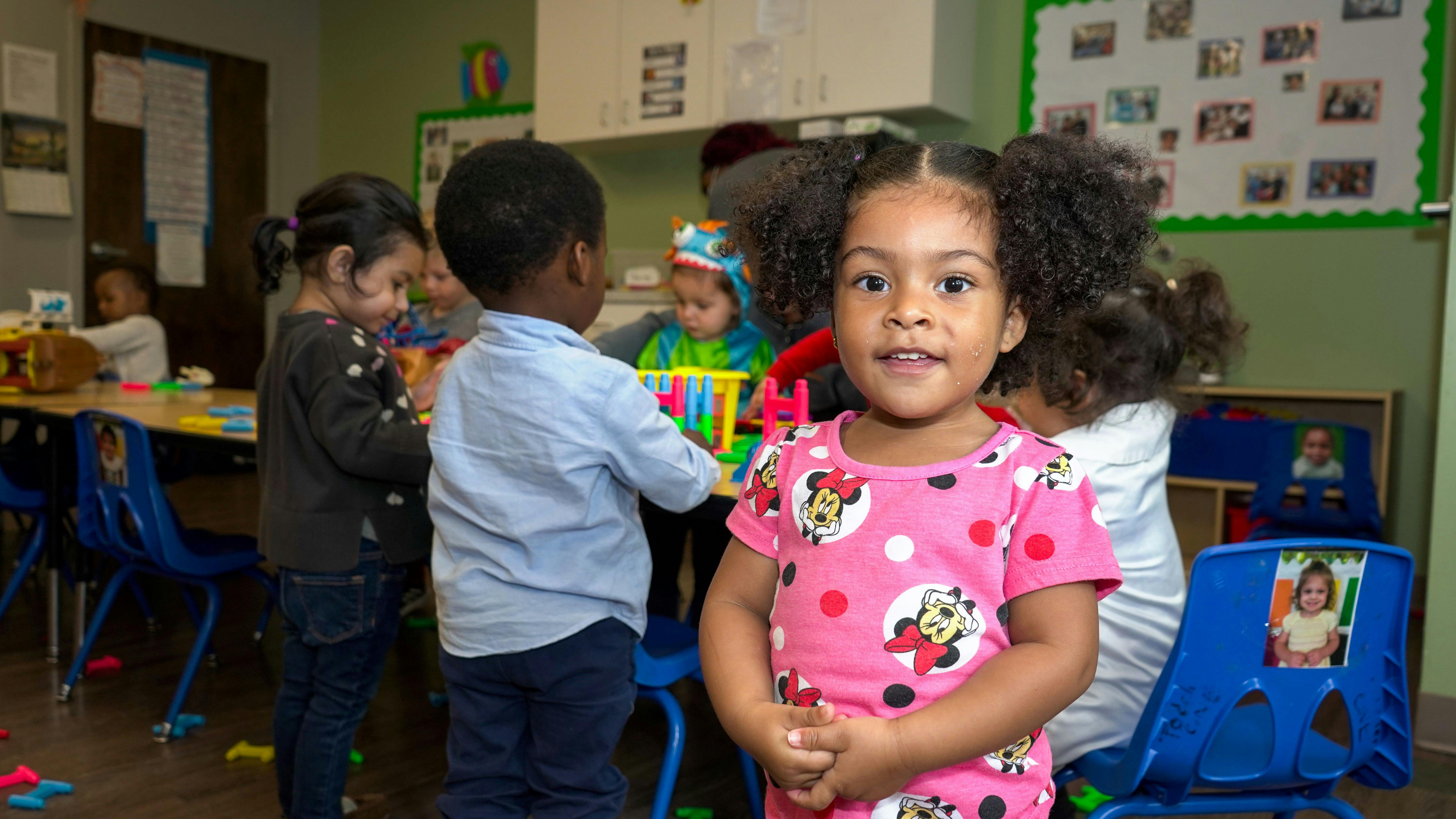 Child in pink shirt standing in a classroom with other children