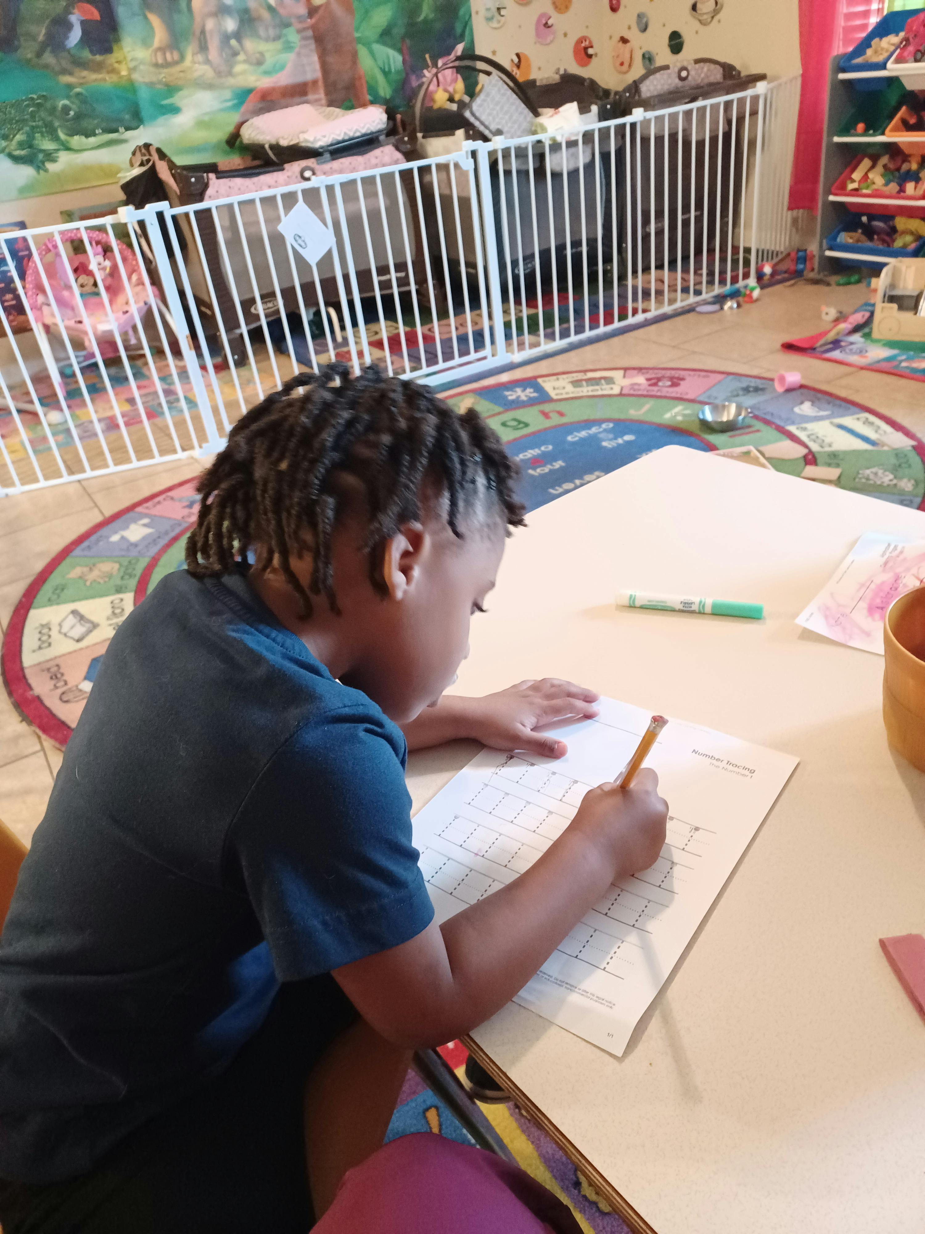 Child writing on paper at a table in a room with a playpen