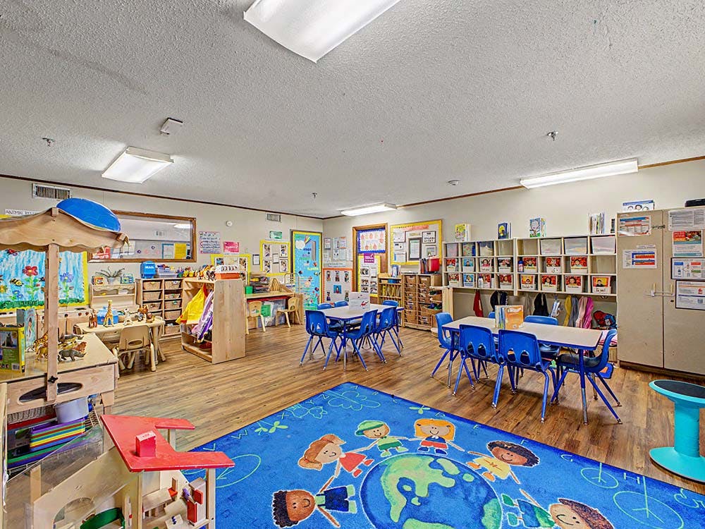 Classroom with small tables and chairs, and a blue rug with children figures