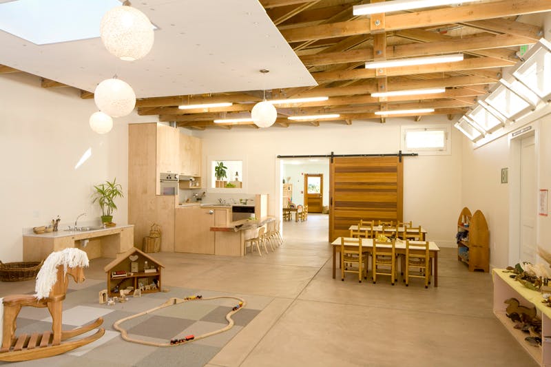 Large room with wooden tables and chairs, a rocking horse, and a dollhouse