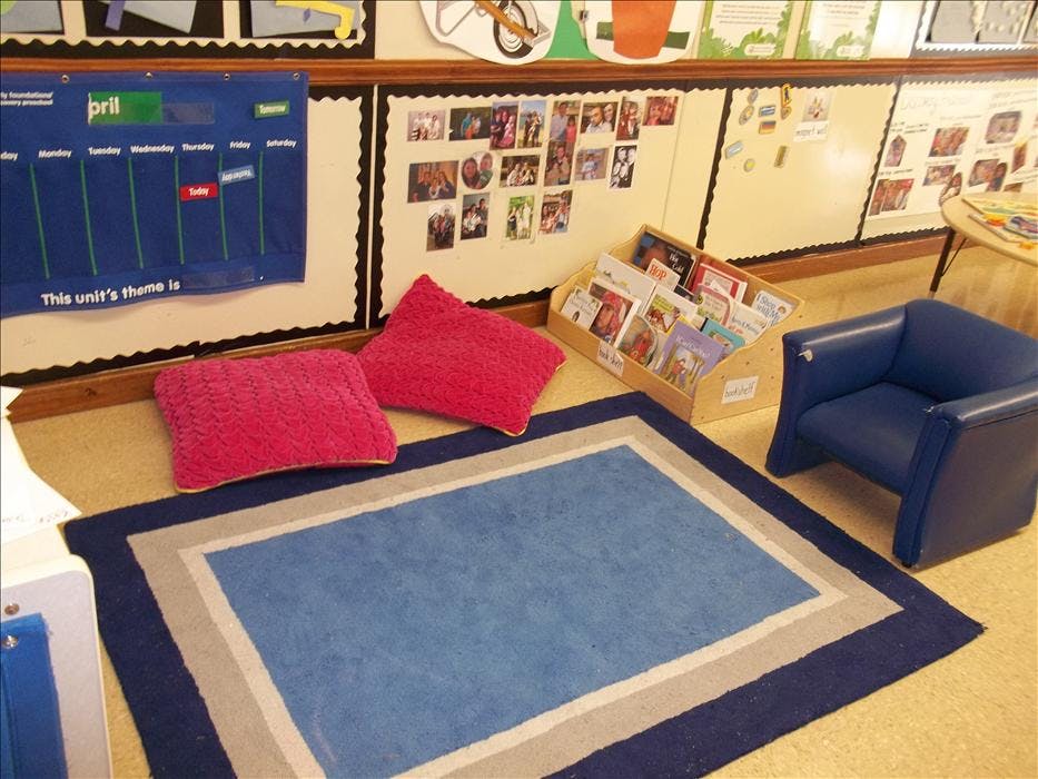 Classroom reading corner with blue rug, cushions, wooden bookshelf, and blue chair