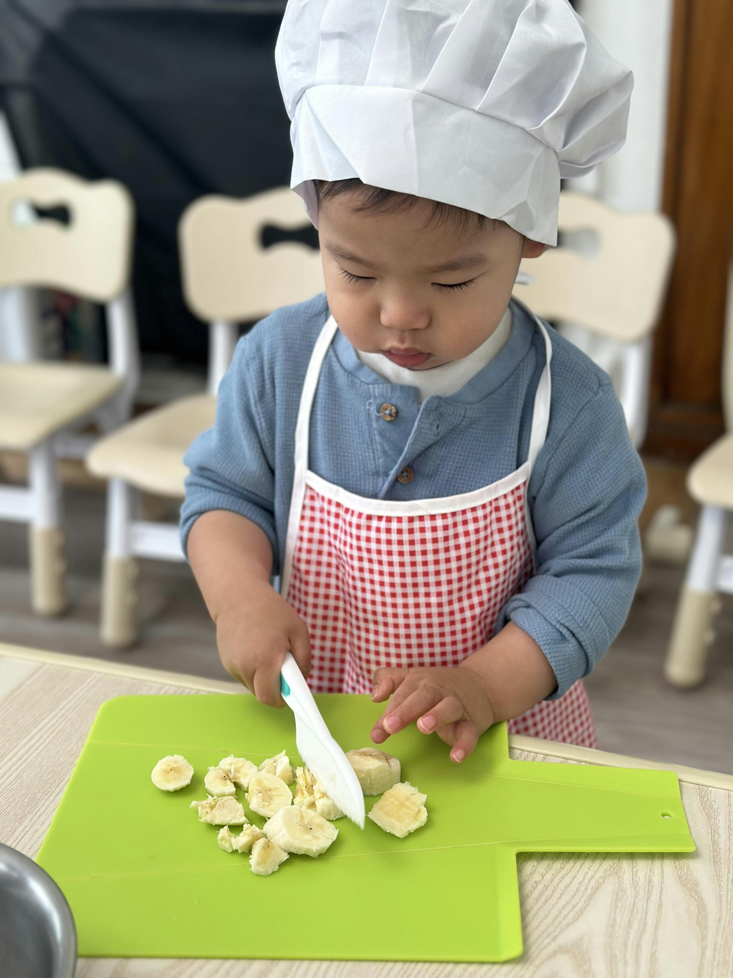 Child wearing a chef hat and apron cutting a banana on a green cutting board