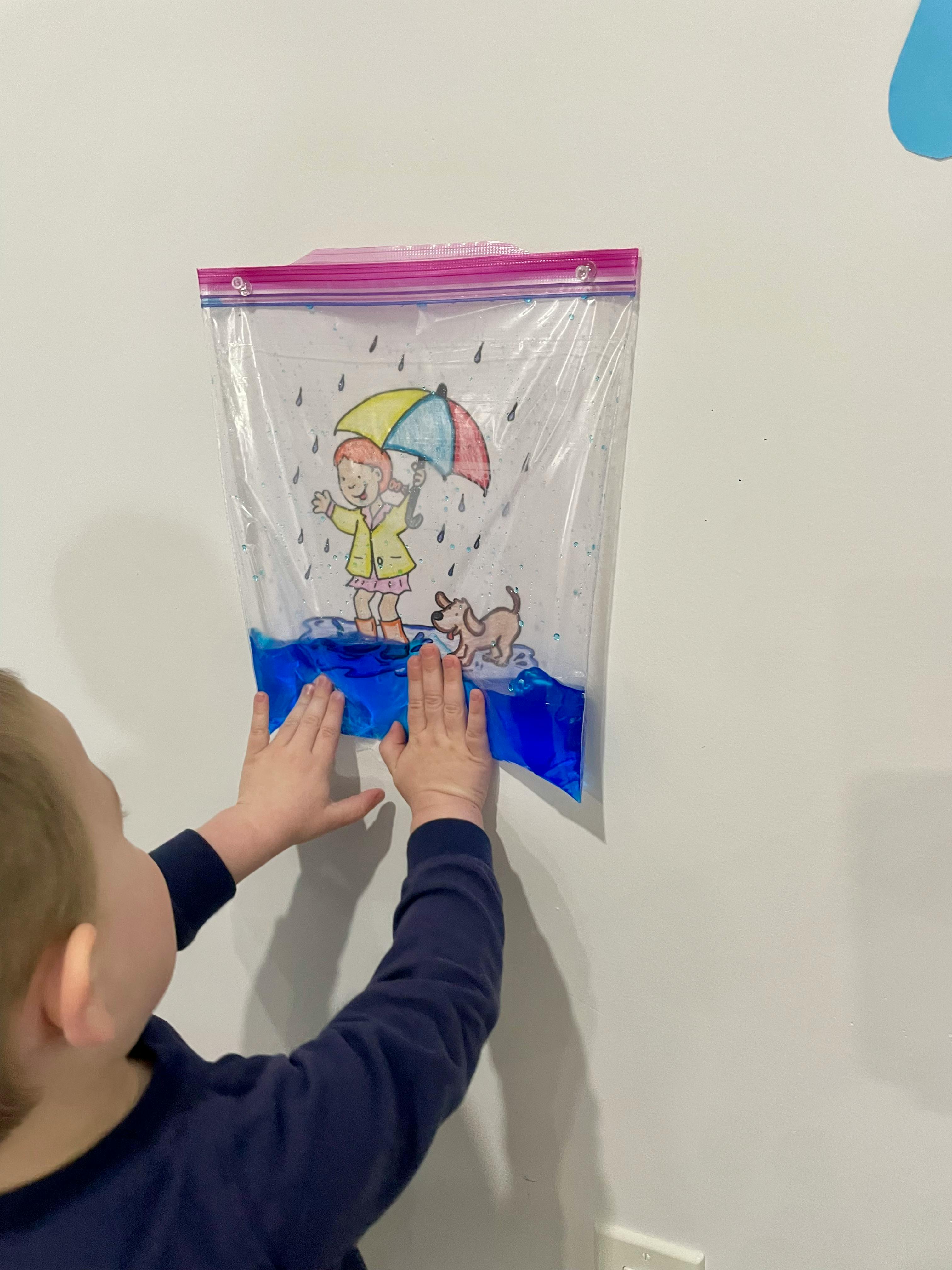 Child pressing hands into a plastic bag with blue liquid and a drawing of a person with an umbrella