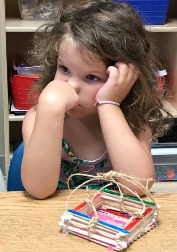 Child with hand on chin looking at a craft made of popsicle sticks