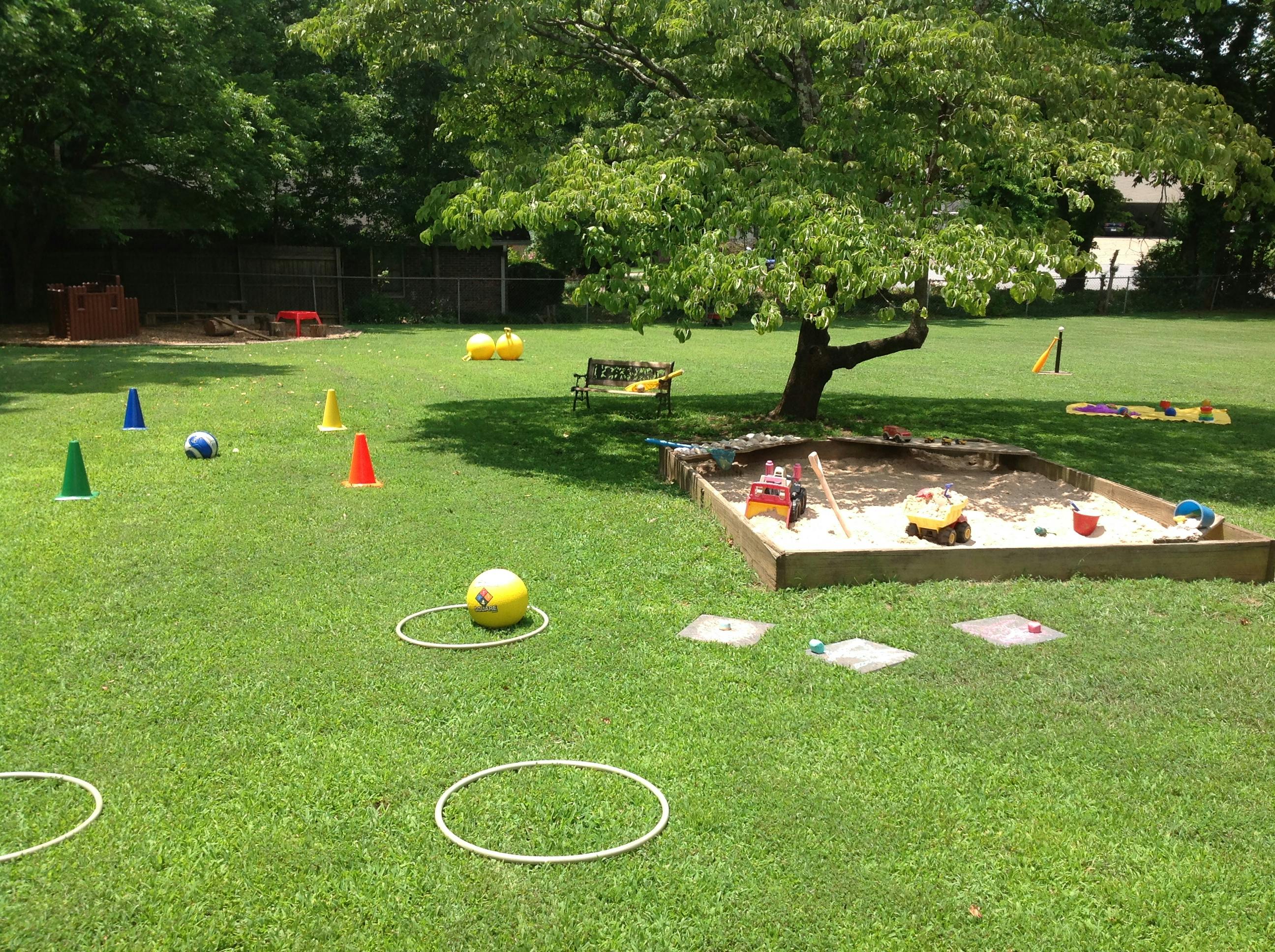 Outdoor play area with sandbox, cones, balls, and hoops on grass