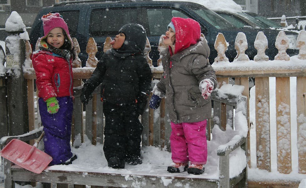 Three children standing in snow next to a wooden fence