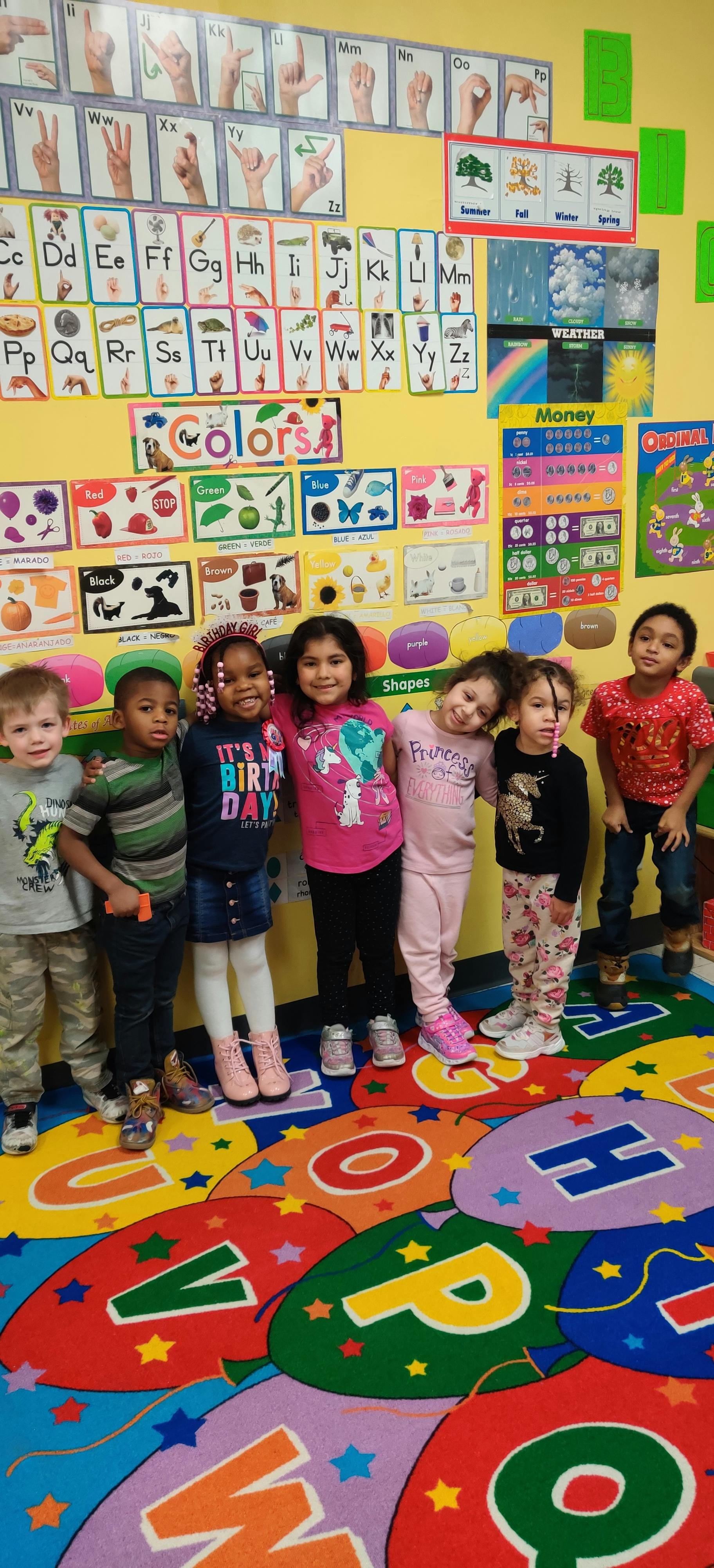 Seven children standing in a classroom with educational posters on the wall