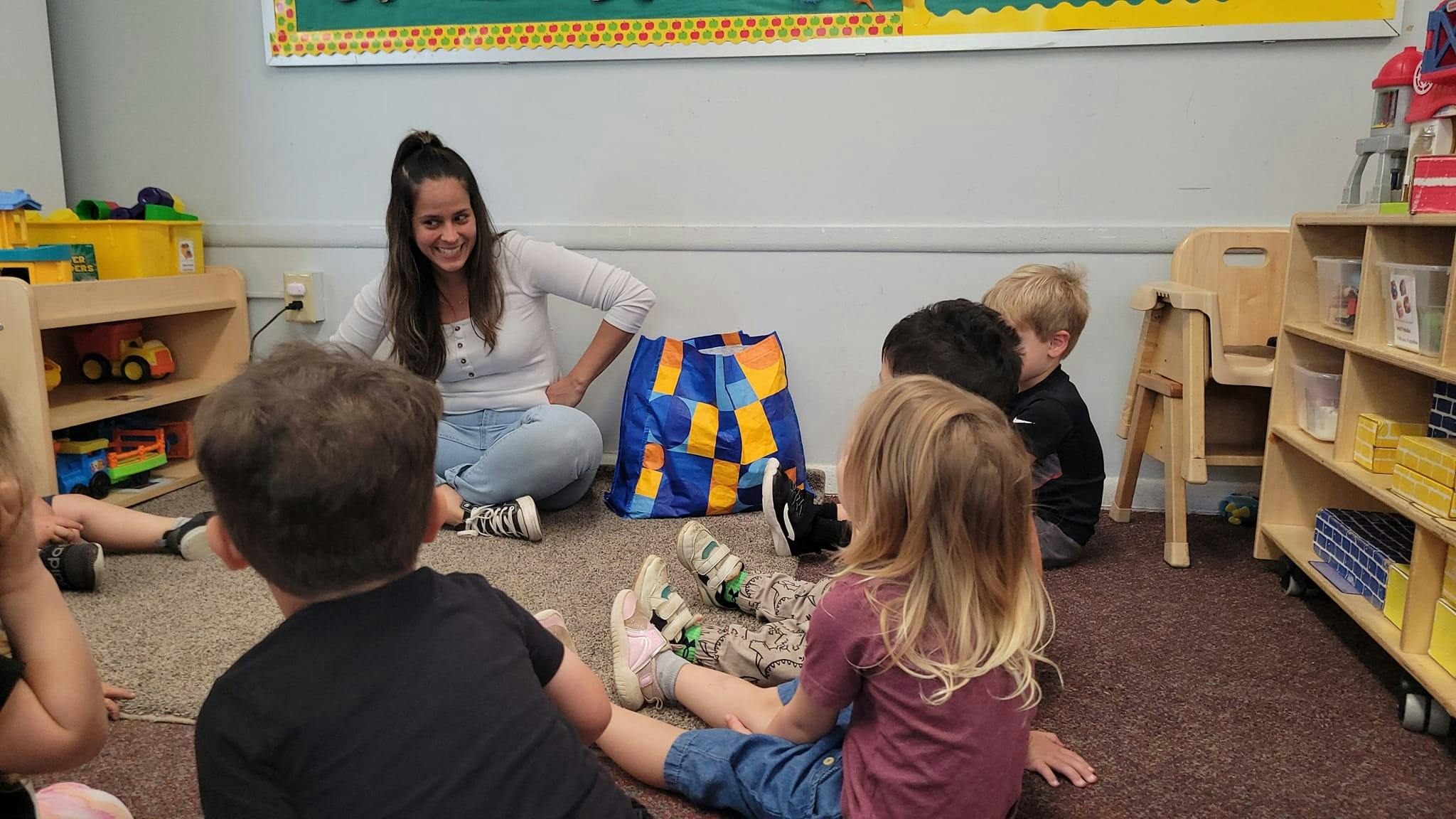 Adult sitting on floor with young children around a large colorful bag in a classroom