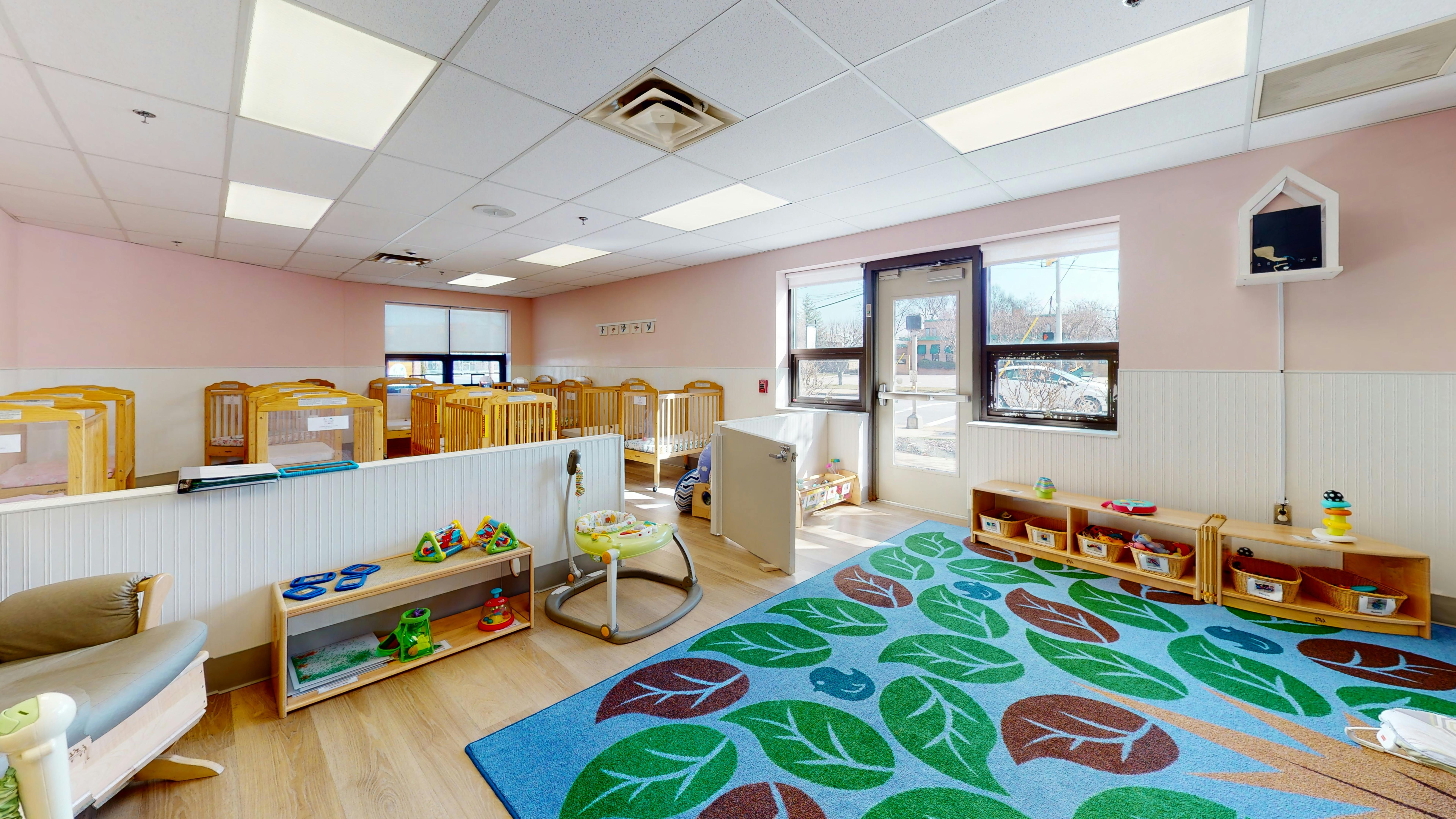 Daycare room with cribs, a colorful rug, and shelves with toys