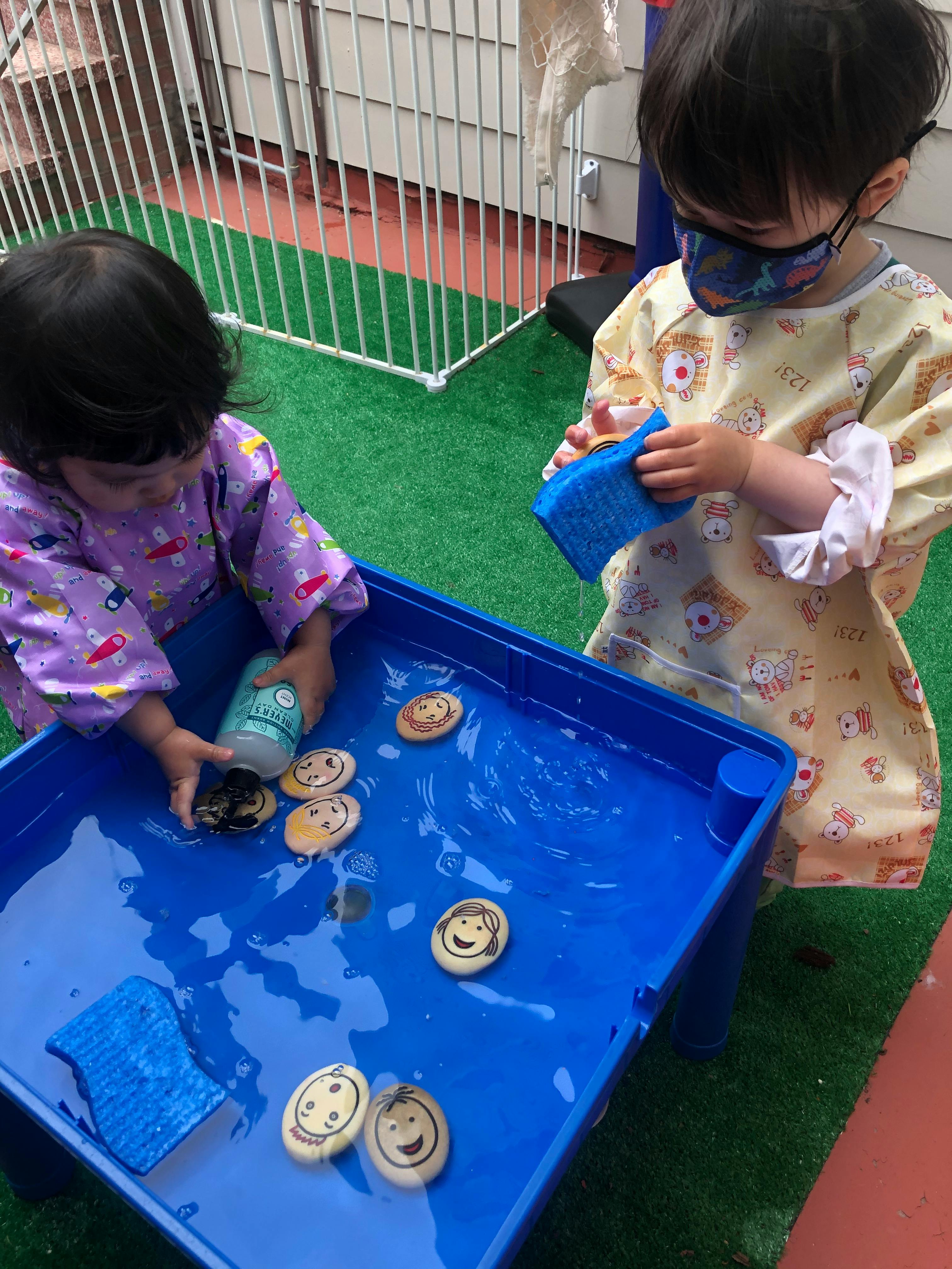 Two children playing with water and painted rocks in a blue bin