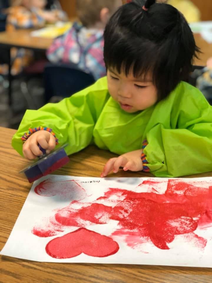 Child in a green smock painting red hearts on paper