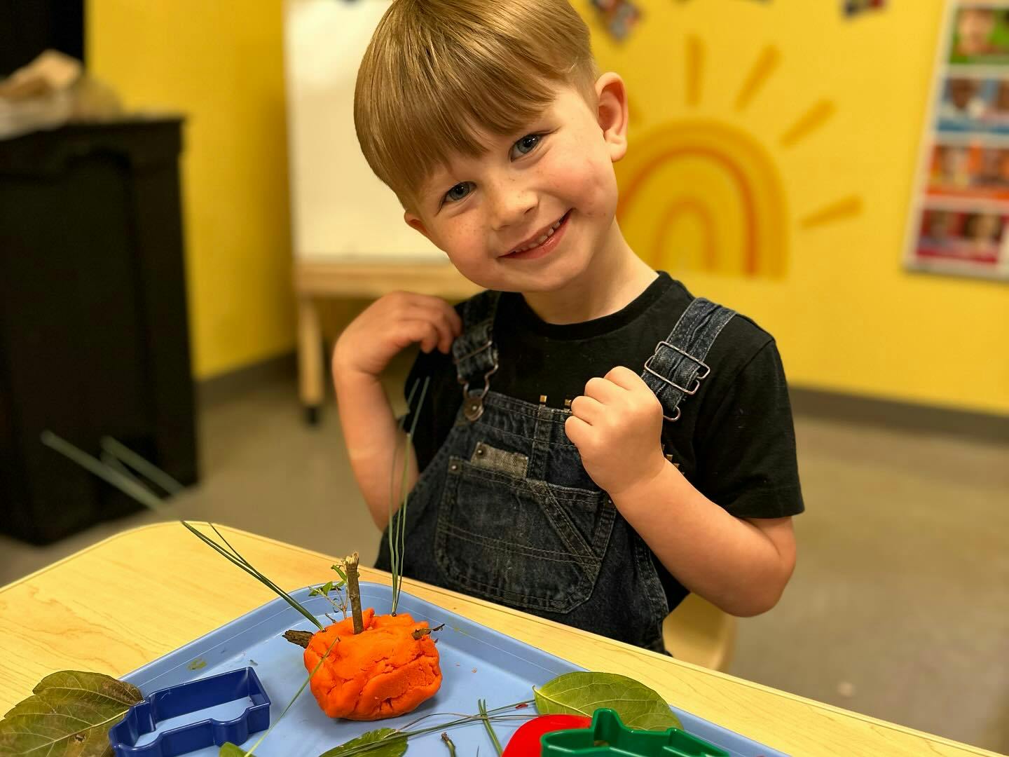 Child in overalls sitting at a table with craft supplies