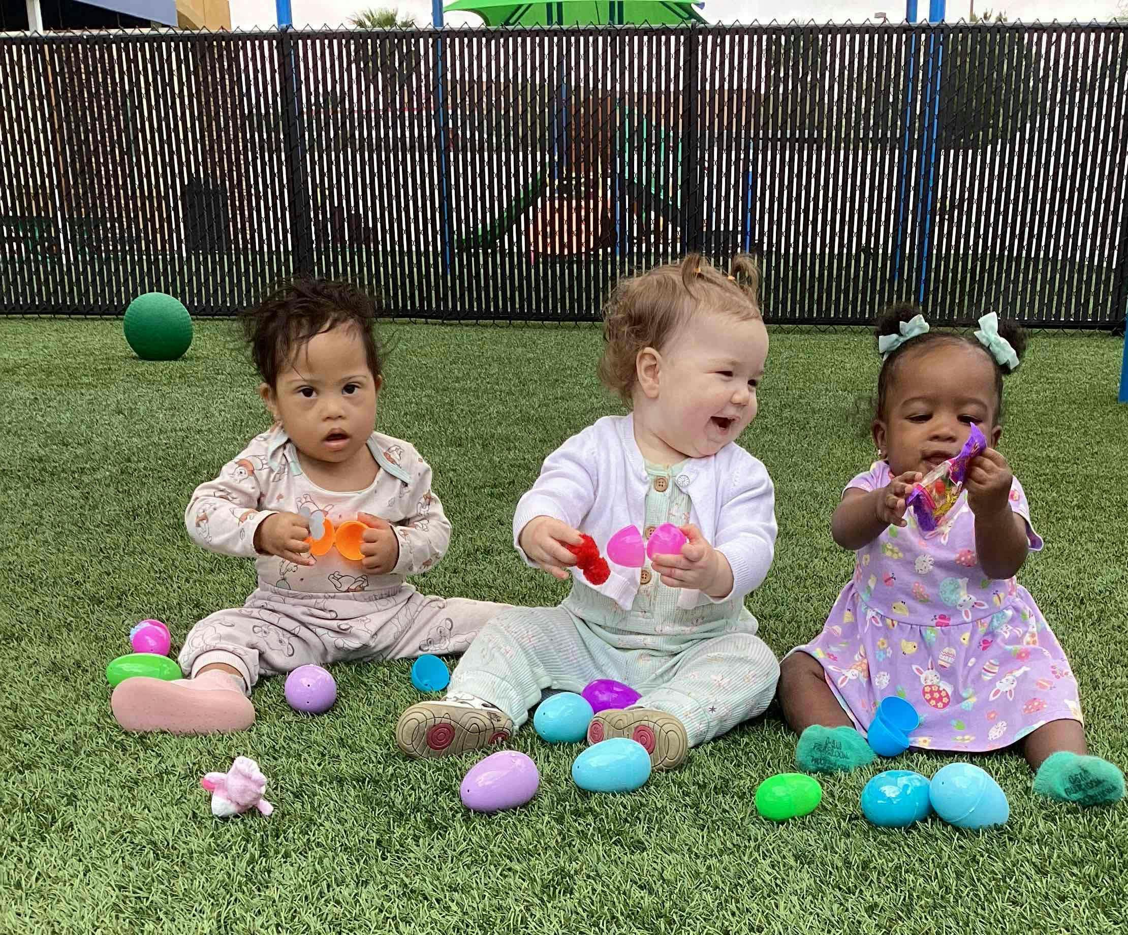 Three children sitting on grass playing with colorful plastic eggs
